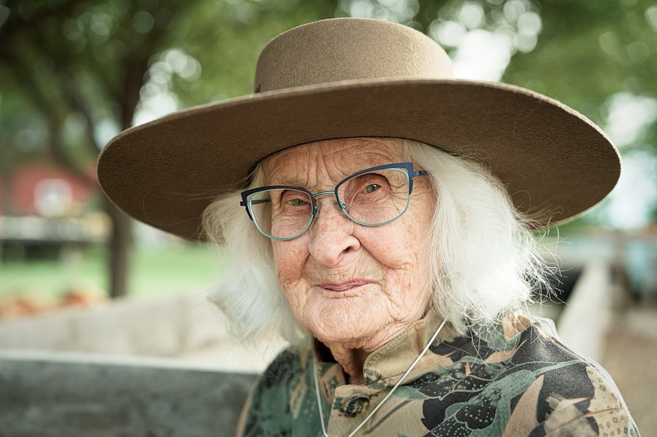 An elderly woman with white hair, wearing glasses, a large wide-brimmed hat, and a patterned shirt outdoors, smiling gently.