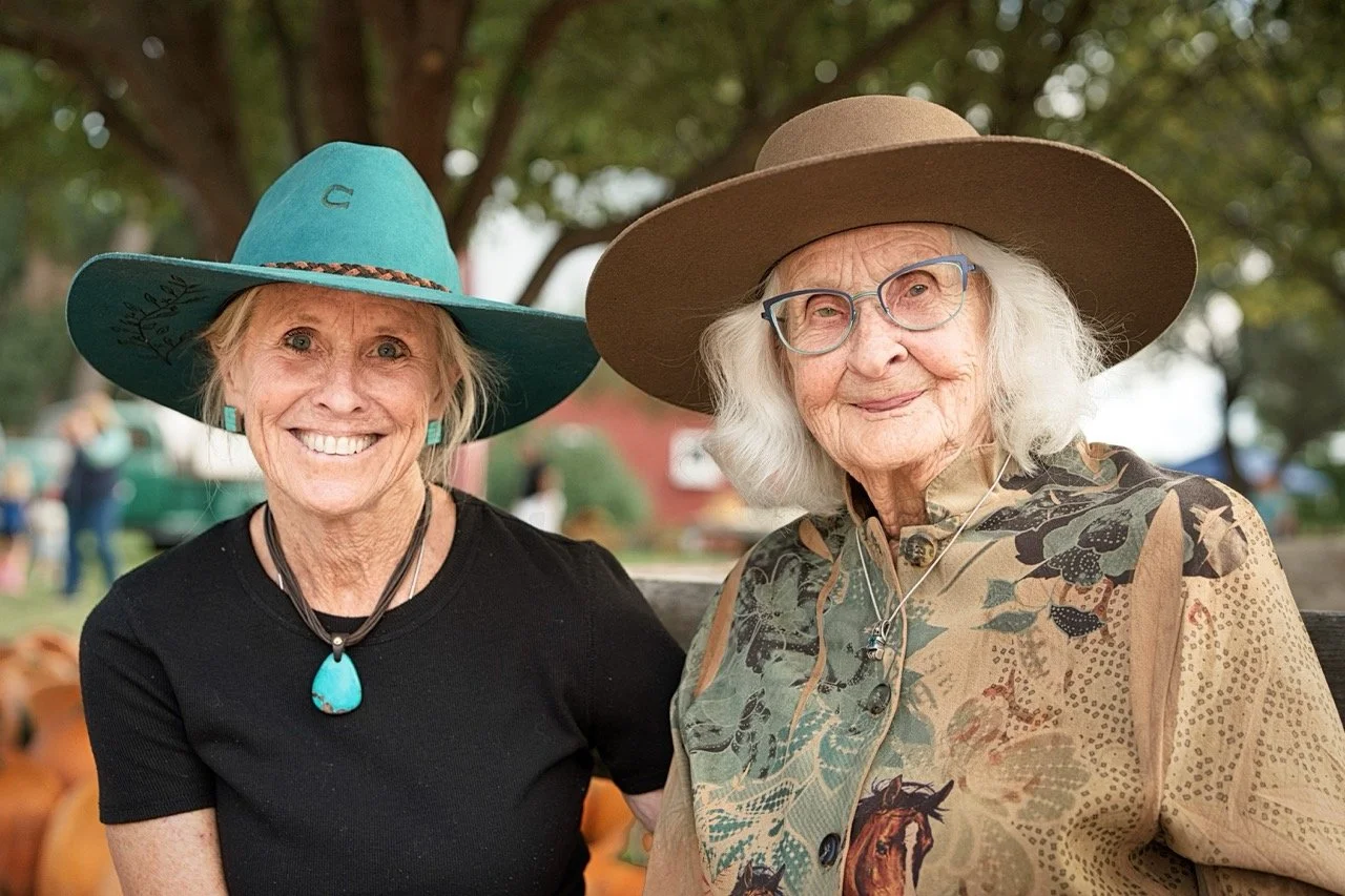 Two elderly women smiling outdoors, wearing large hats and colorful clothing, with trees and pumpkins in the background.