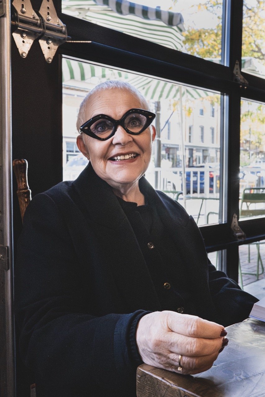 An elderly woman with short white hair and large black cat-eye glasses, smiling warmly, seated at a table in a cafe. She is wearing a dark jacket and is holding something in her hand. Behind her, large windows reveal a street scene with parked cars a