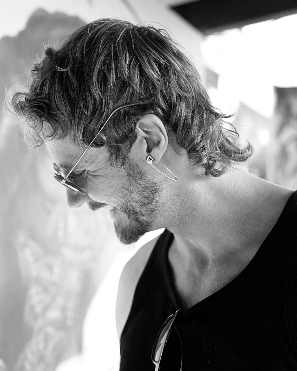 A side profile of a man with wavy hair, glasses, and earphones, smiling while looking down.