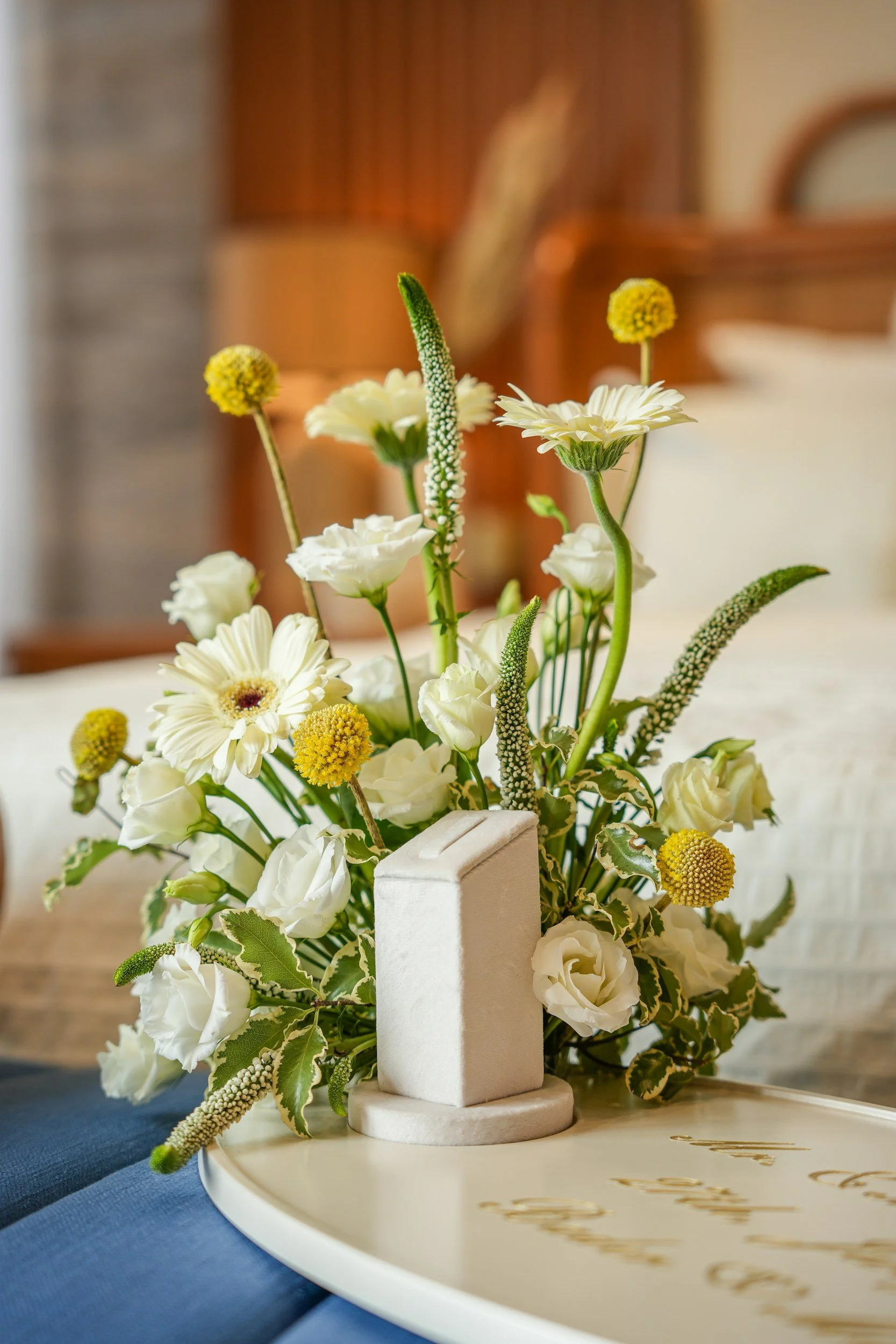 A floral arrangement with white daisies and other white flowers, accented with green and yellow buds, placed in a small white rectangular container on a white tray with gold lettering, on a table with a blue cloth.