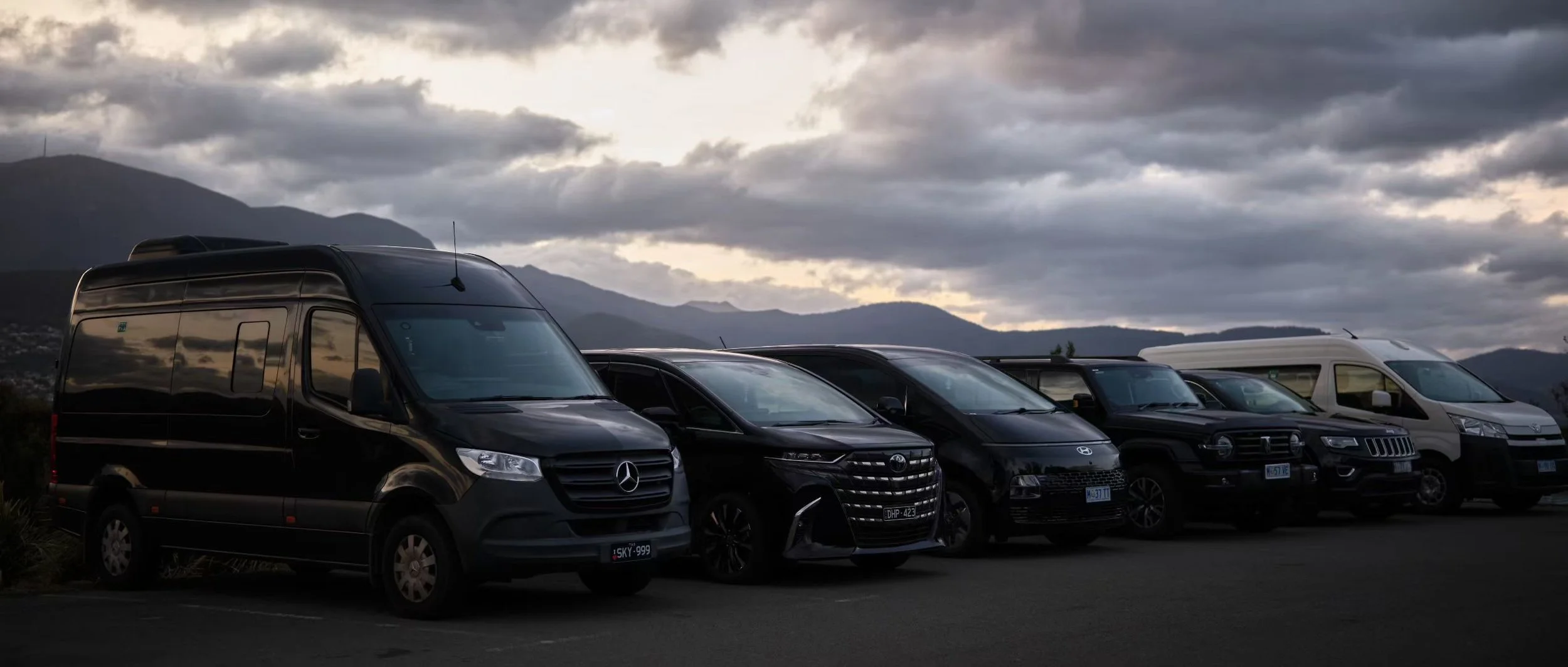 Line of various black and white vans and SUVs parked outdoors with mountain range and cloudy sky in the background.