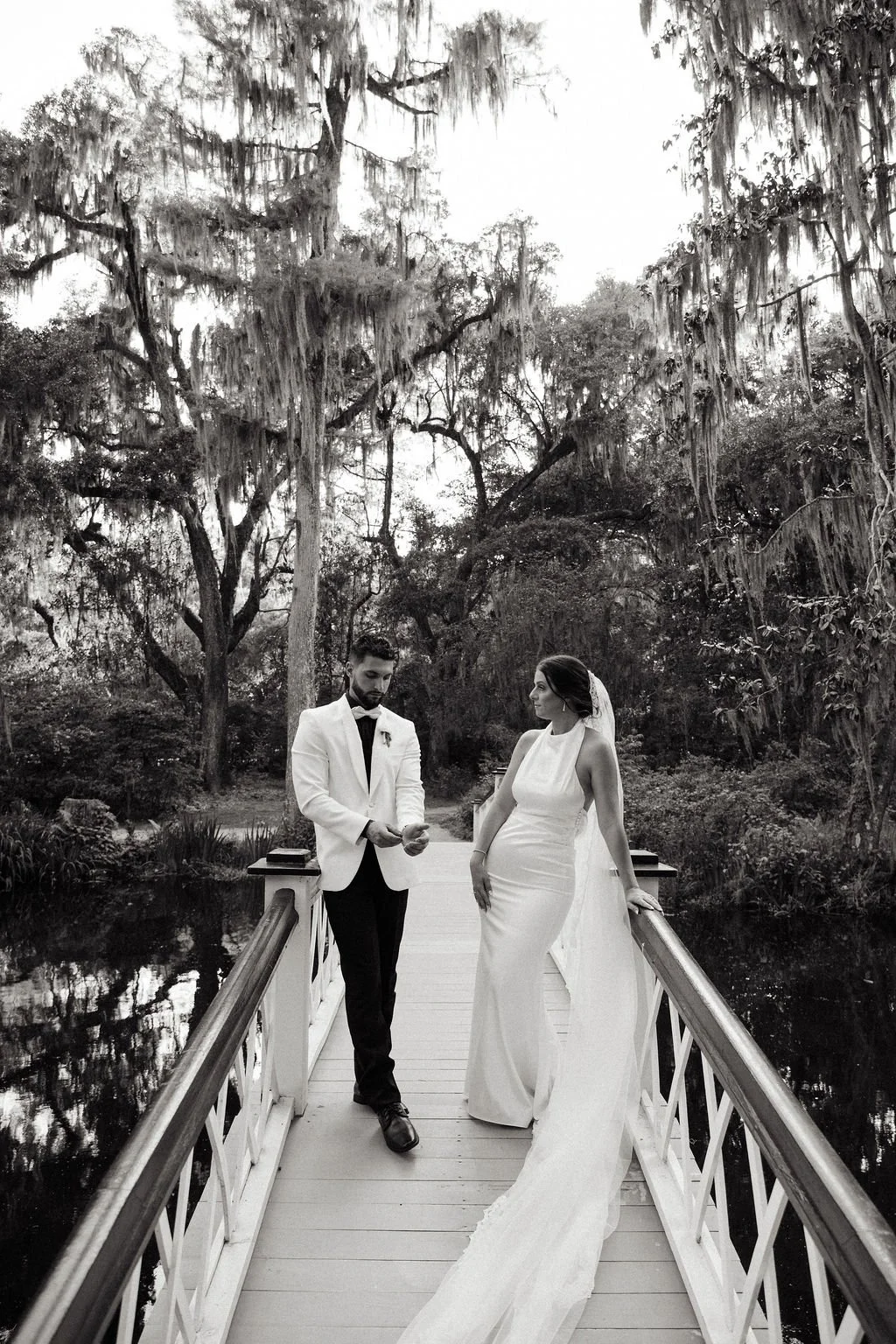 Black and white photo of a bride and groom on a small bridge in a wooded area, with tall hanging moss trees in the background. The groom, wearing a white tuxedo jacket and bow tie, is looking down at his hands, while the bride, in a long elegant whit