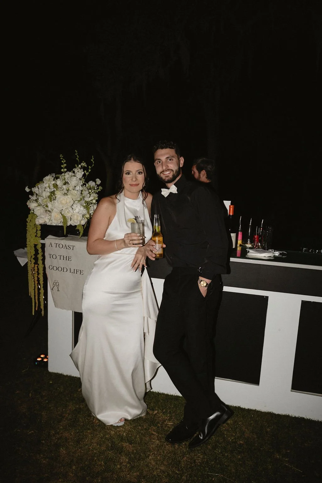 A newlywed couple at a wedding reception, standing in front of a bar, smiling and holding drinks, with a floral arrangement and a sign that reads 'A toast to the good life' in the background.