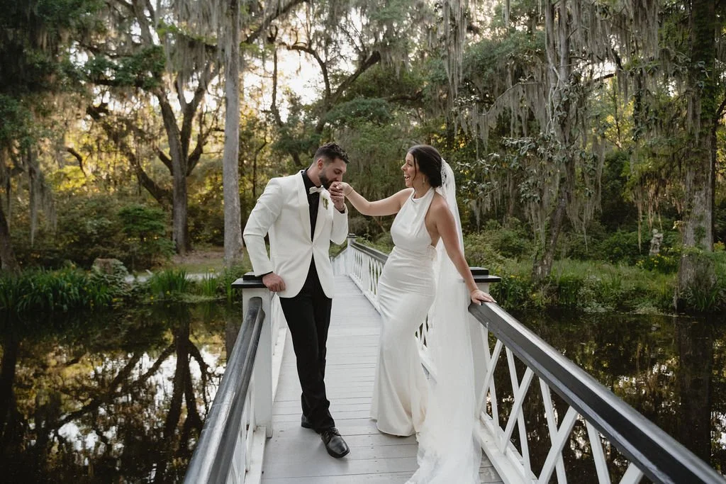 A bride and groom on a white bridge over water surrounded by trees, with the bride touching the groom's face as he smiles.