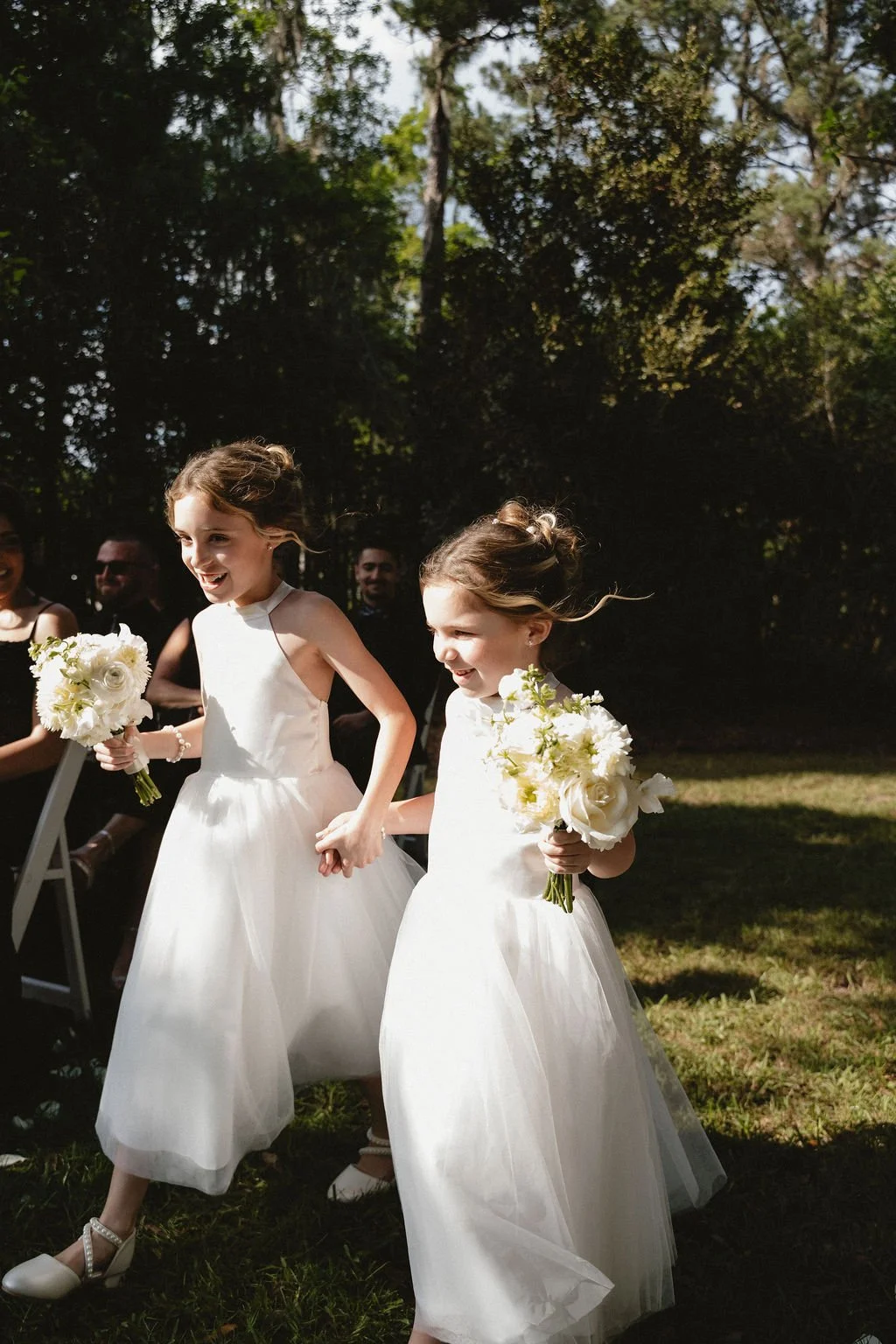 Two young girls in white dresses holding bouquets of flowers, holding hands and walking outdoors during a wedding ceremony, with adults and trees in the background.