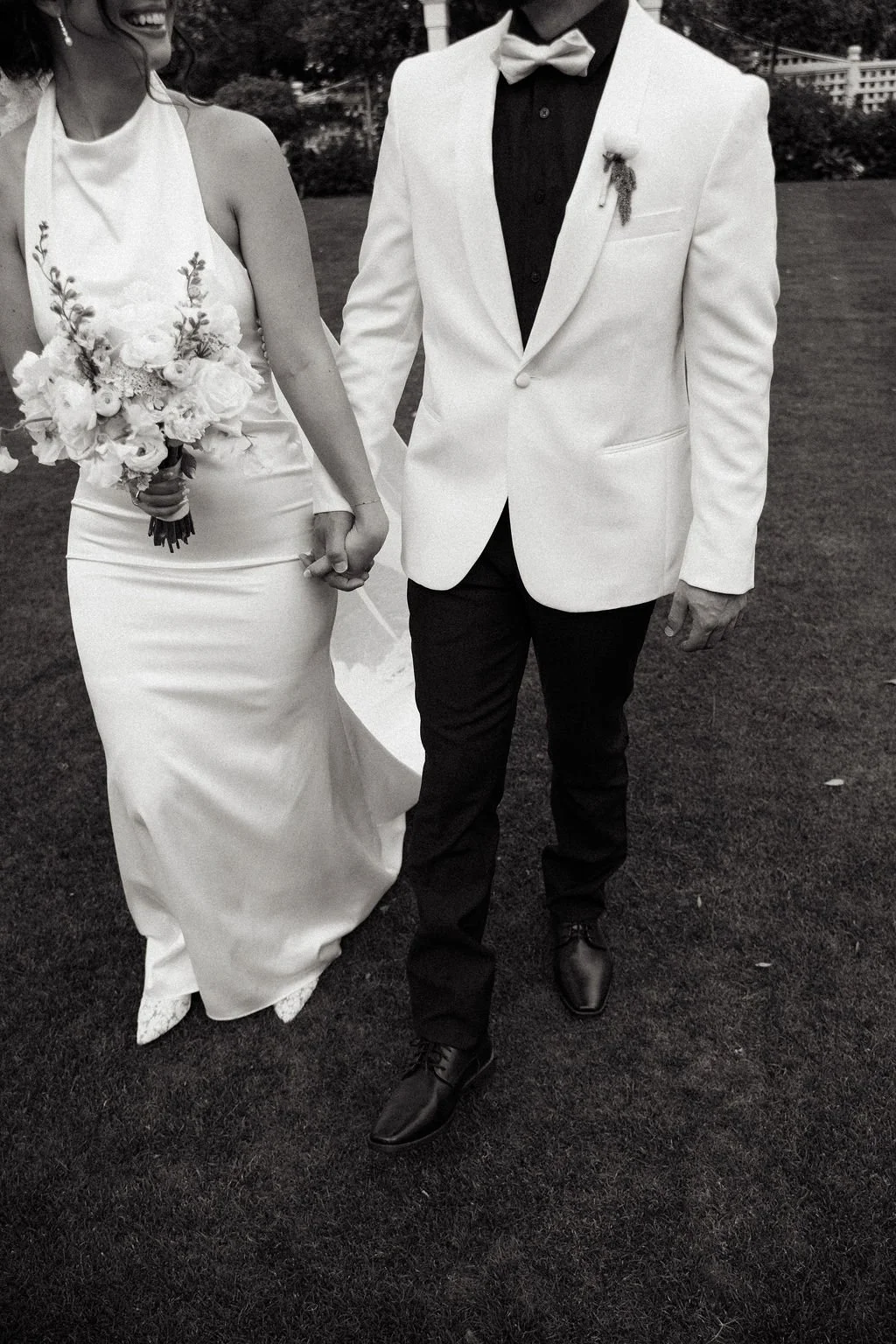 A bride and groom holding hands walking outdoors on a grassy area, wedding attire with the bride in a white gown and the groom in a white jacket and black pants, holding a bouquet of flowers.
