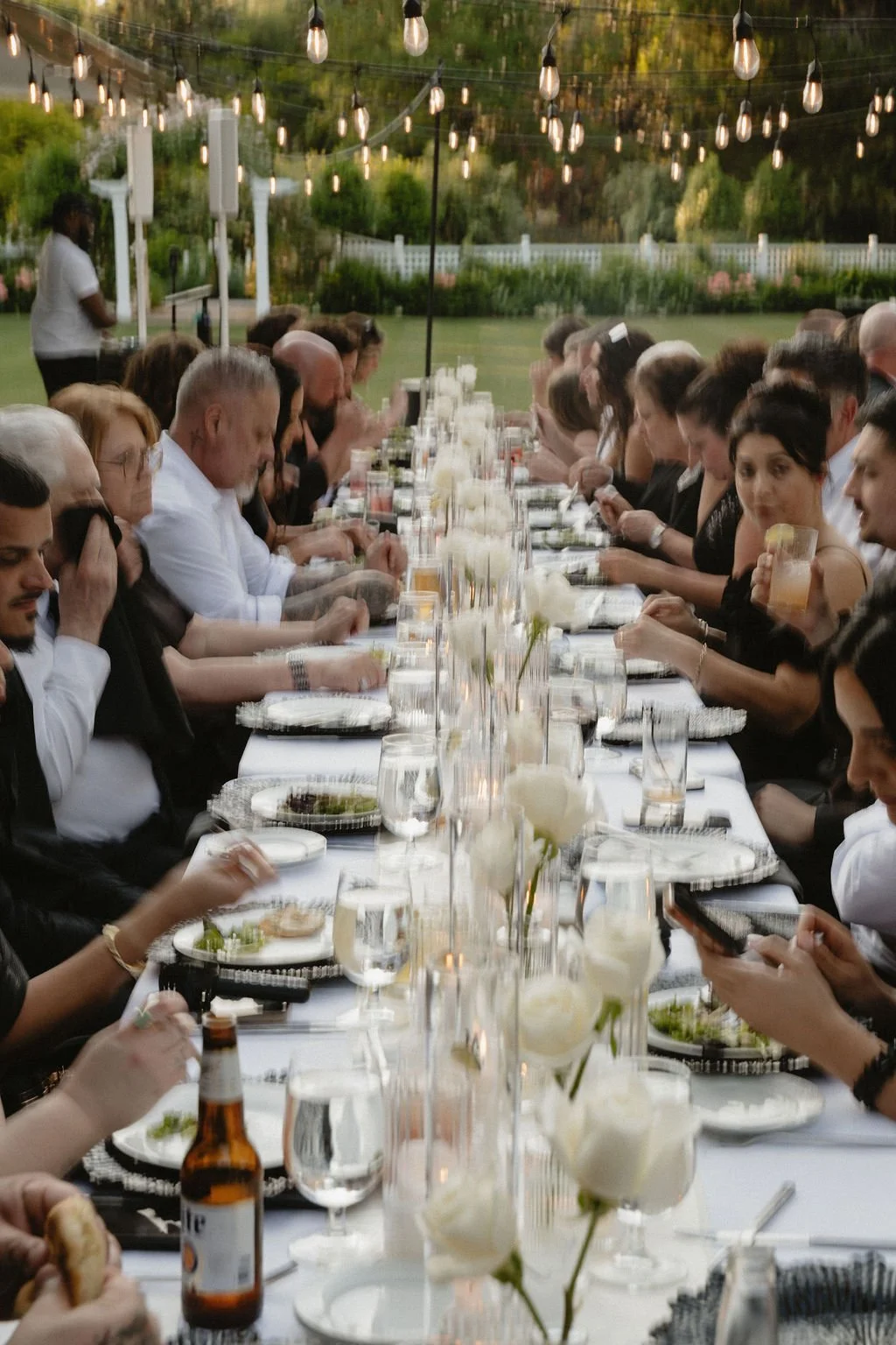 Long outdoor dinner table with many people dressed in formal attire, decorated with white flowers and candles, under string lights, in a lush garden setting.