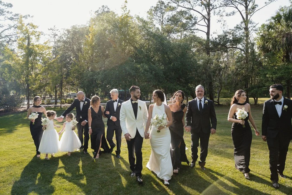 Wedding party walking on a grassy field in a park during daytime, with trees in the background. The bride in a white gown and bridesmaids in black dresses hold bouquets, while the groom and groomsmen wear suits with bow ties.