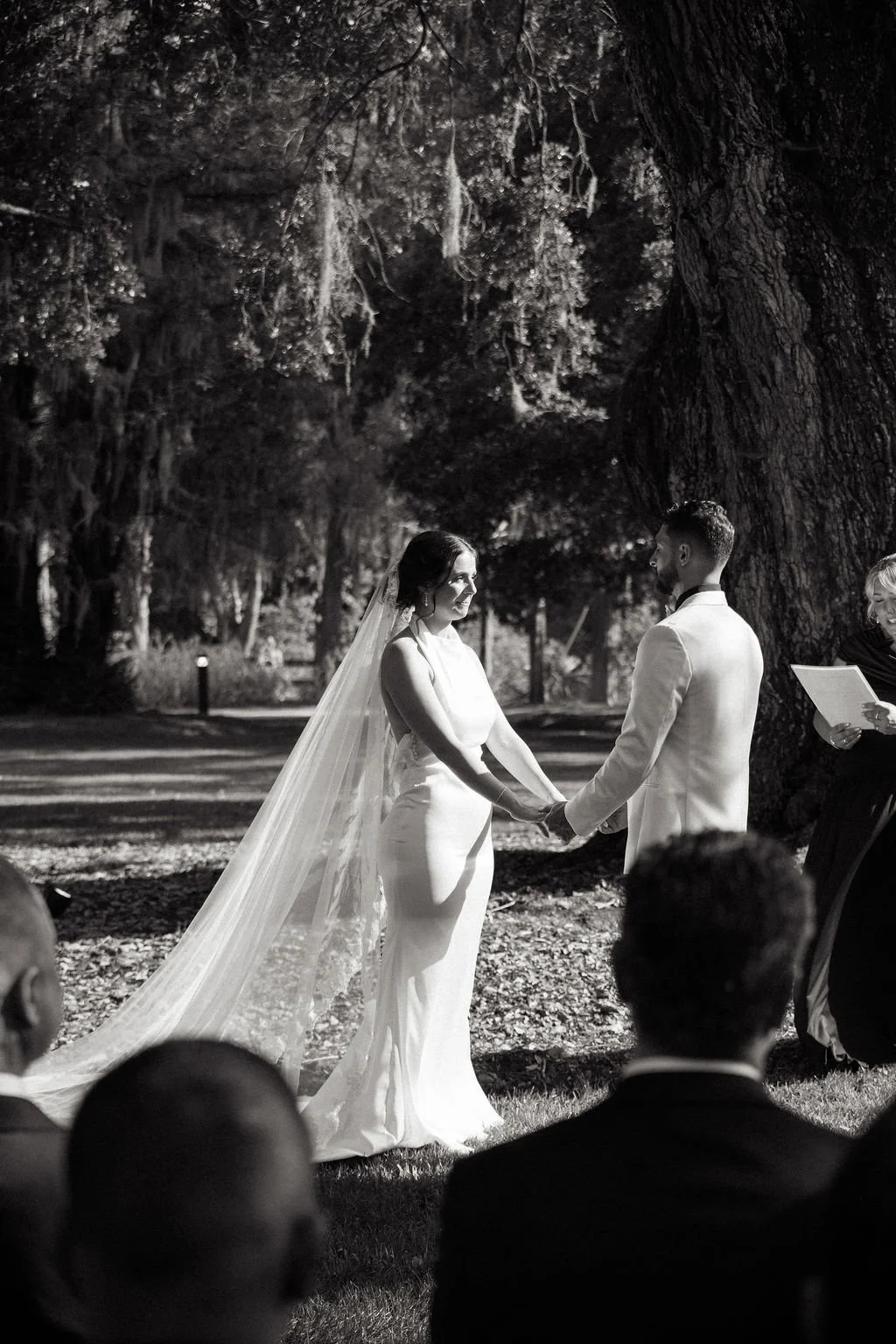 A black and white photo of a wedding ceremony outdoors at night, with a bride and groom holding hands and exchanging vows under a large tree, surrounded by guests and an officiant.