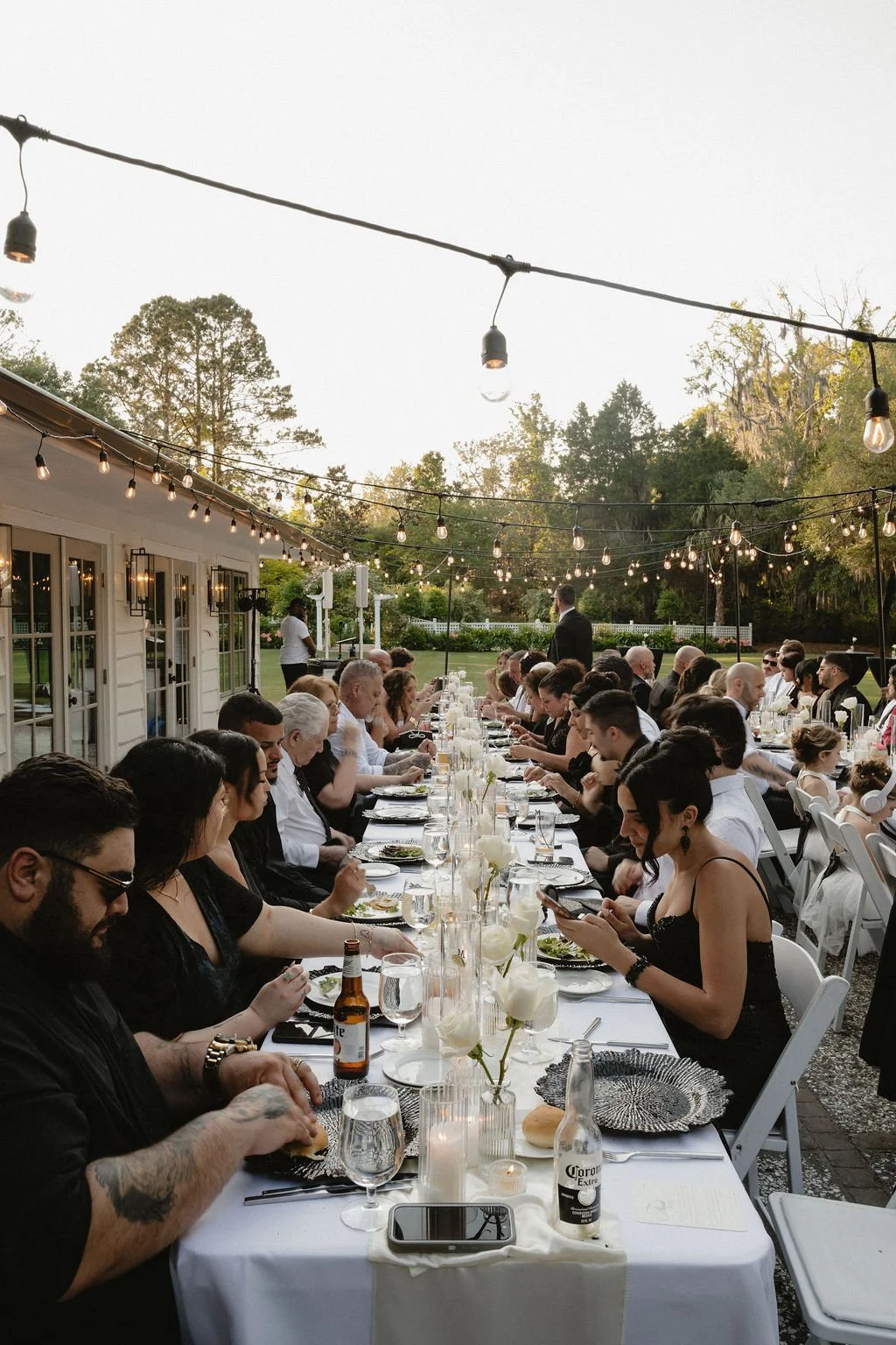 People dining at a long outdoor table during sunset with string lights and green trees in the background.