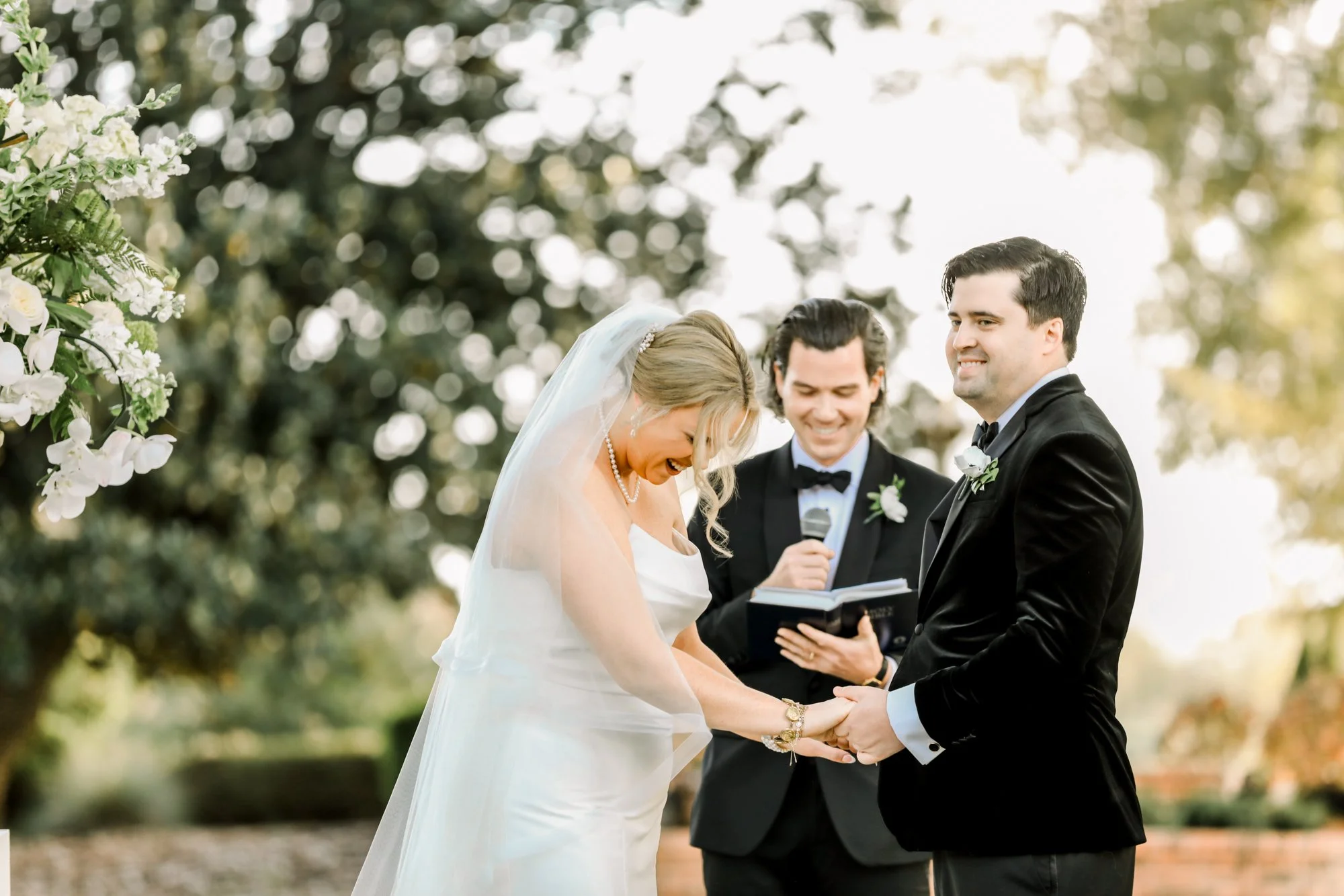 A wedding ceremony taking place outdoors with a bride and groom holding hands, someone officiating reading from a book, a woman with a veil and pearl necklace smiling, and a man in a black tuxedo smiling among trees and bright sunlight.