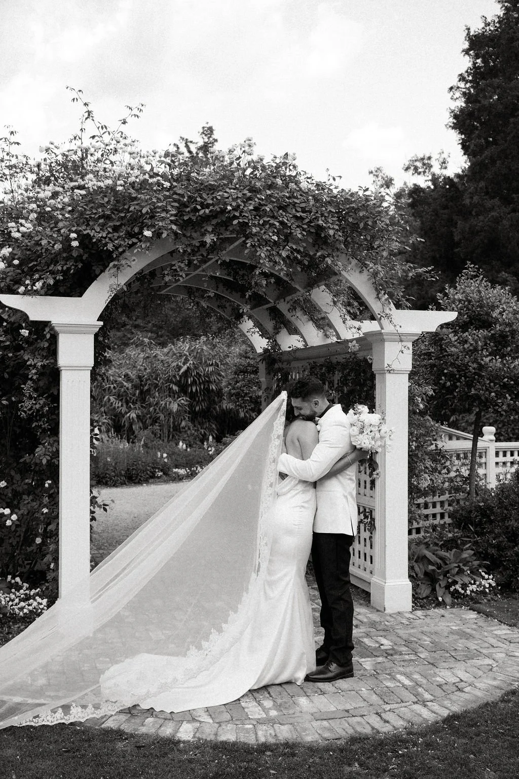 A bride and groom embrace under a floral archway during their wedding in an outdoor garden setting, with greenery and trees in the background.