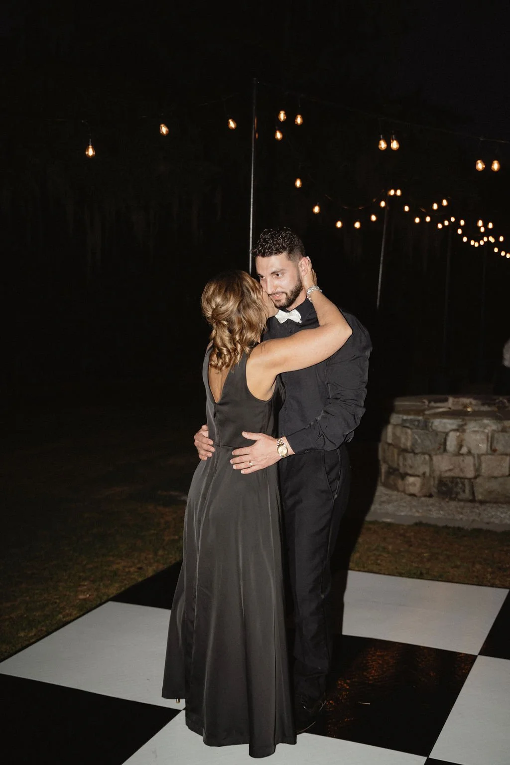 A couple dancing at a nighttime event with string lights hanging above. The woman is wearing a black sleeveless dress, and the man is dressed in a black shirt and black trousers. They are embracing on a black-and-white checkered dance floor.