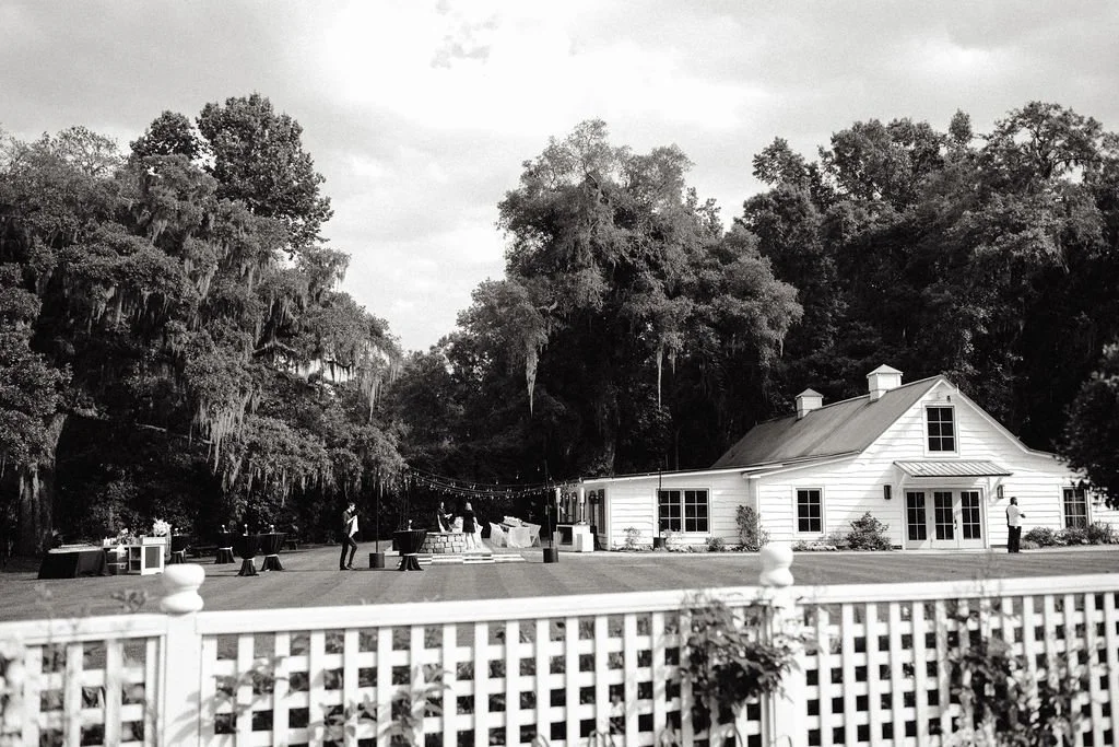 Black and white photo of an outdoor event on a lawn with a white house, trees, and a fence in the foreground.