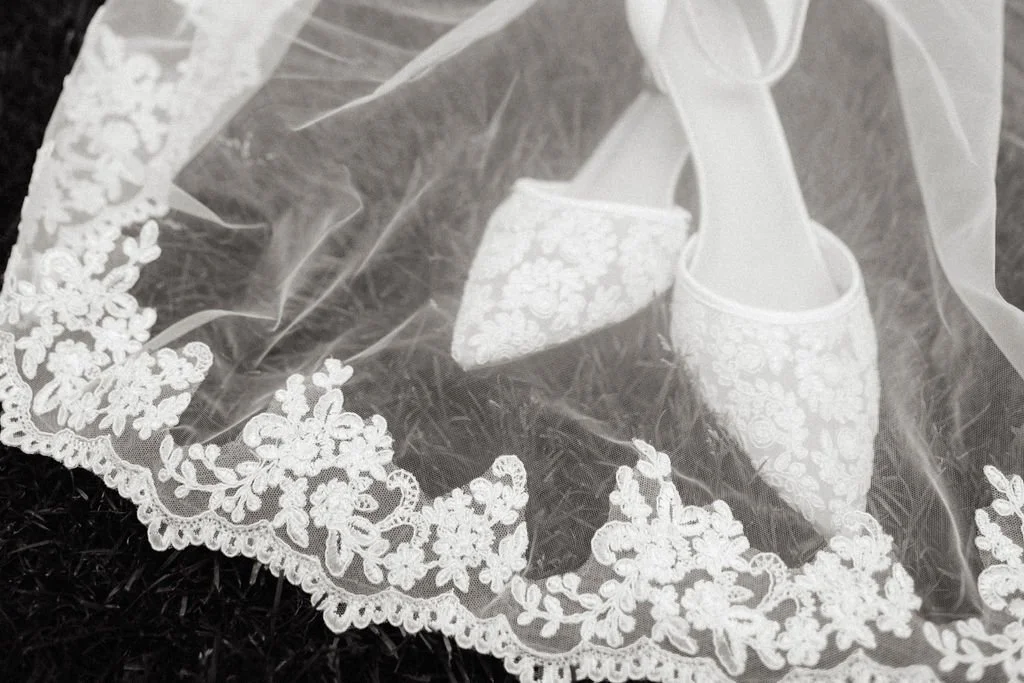 A pair of white embroidered shoes on lace fabric with floral pattern and scalloped edge.