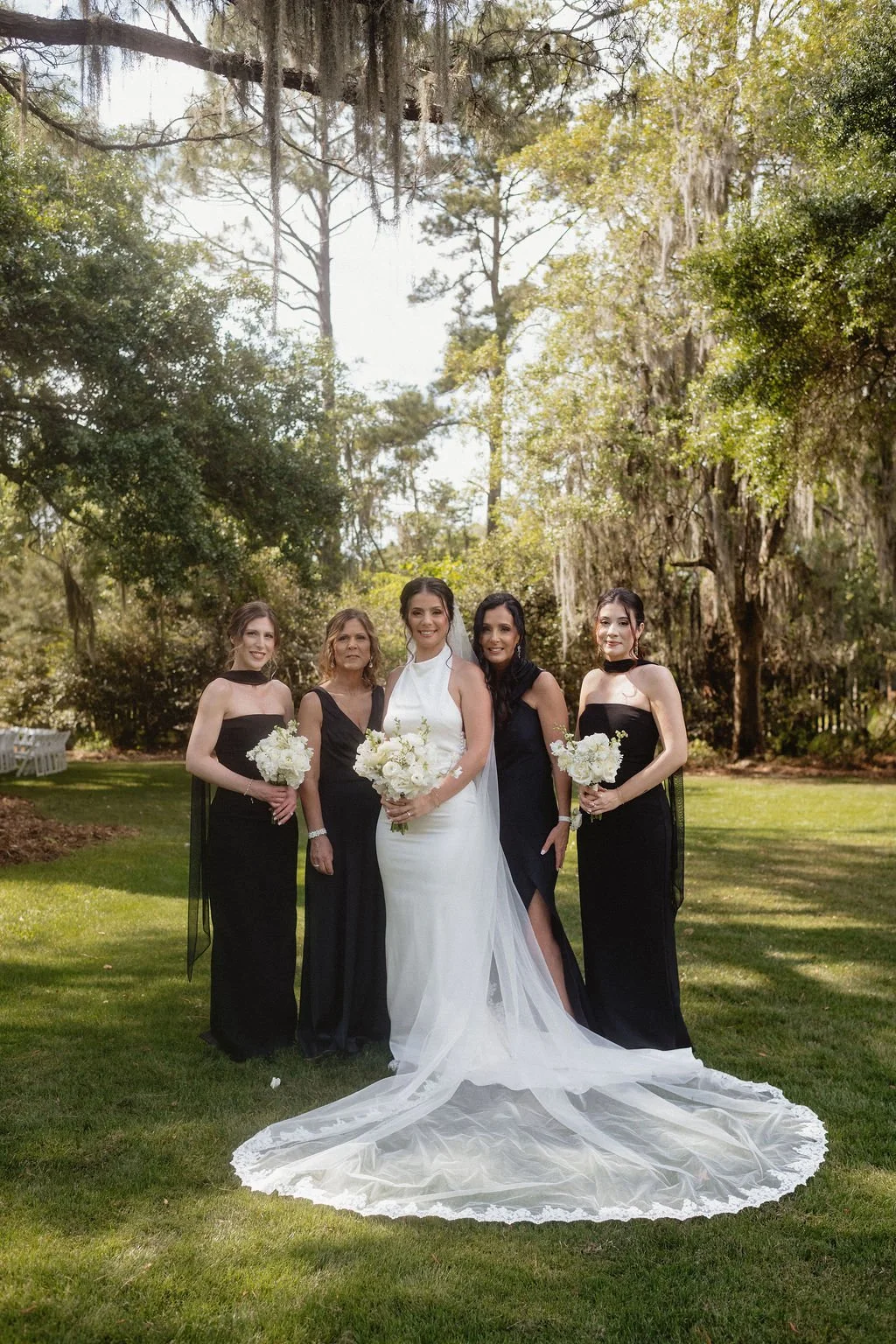 Five women dressed for a wedding, standing outdoors on a lush green lawn with trees and Spanish moss hanging from branches in the background. The woman in the center is in a white wedding gown, holding a bouquet of white flowers. The other four women