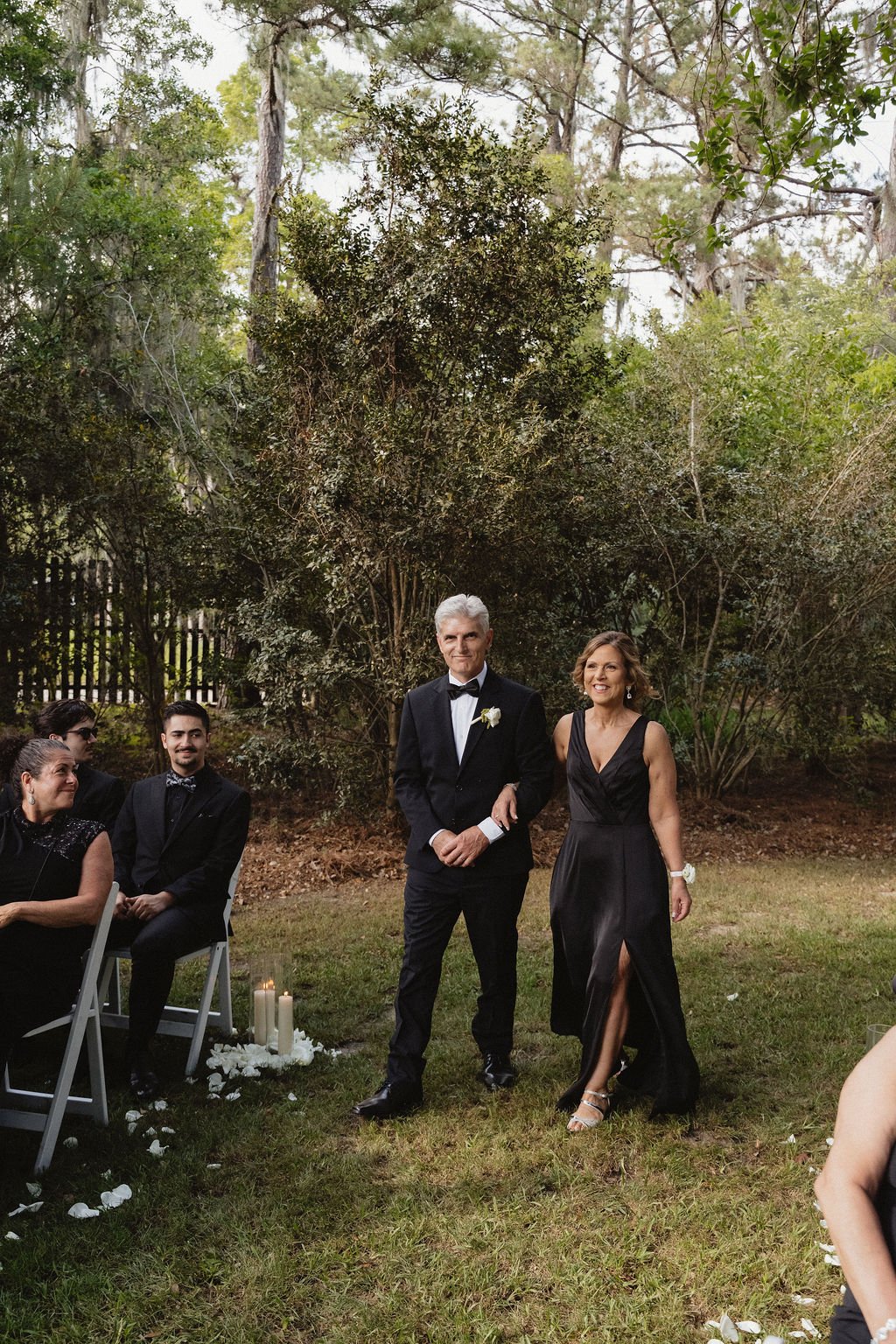 A couple dressed in formal attire walking outdoors at a wedding ceremony, with guests seated nearby in a garden setting.