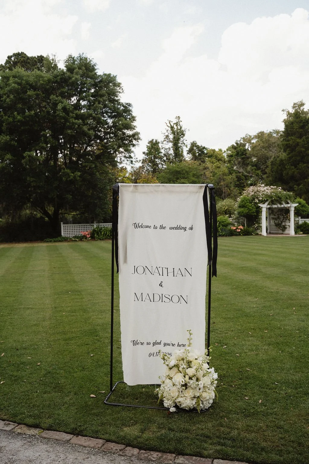 A wedding sign outdoors on a grassy lawn, welcoming guests to the wedding of Jonathan and Madison, decorated with a white floral arrangement at the base.