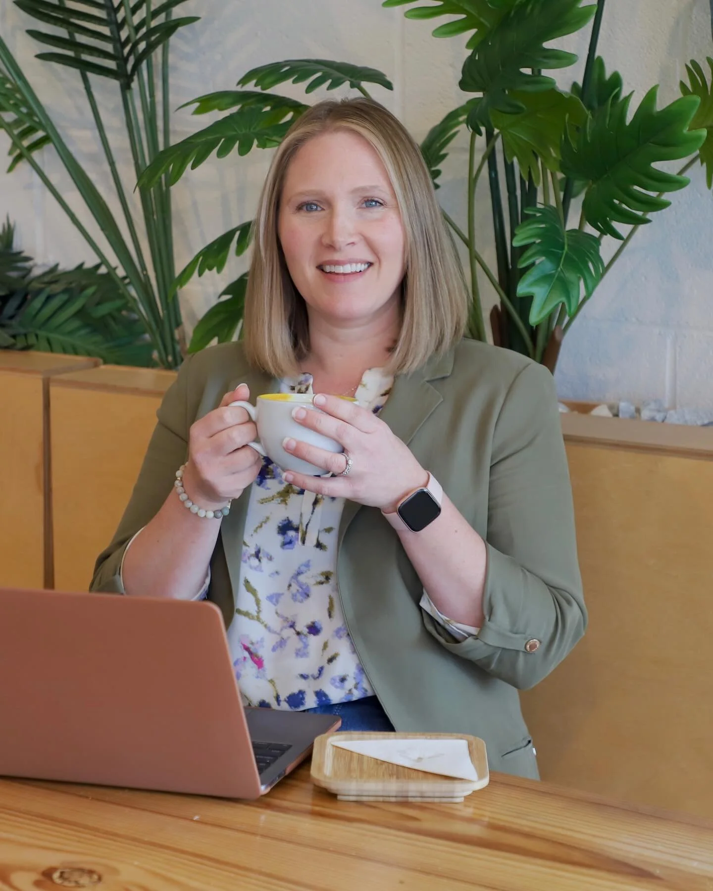 A woman with shoulder-length blonde hair, smiling, holding a white mug, sitting at a wooden table with a pink laptop and wooden tray, in a room with large green indoor plants and a white brick wall.