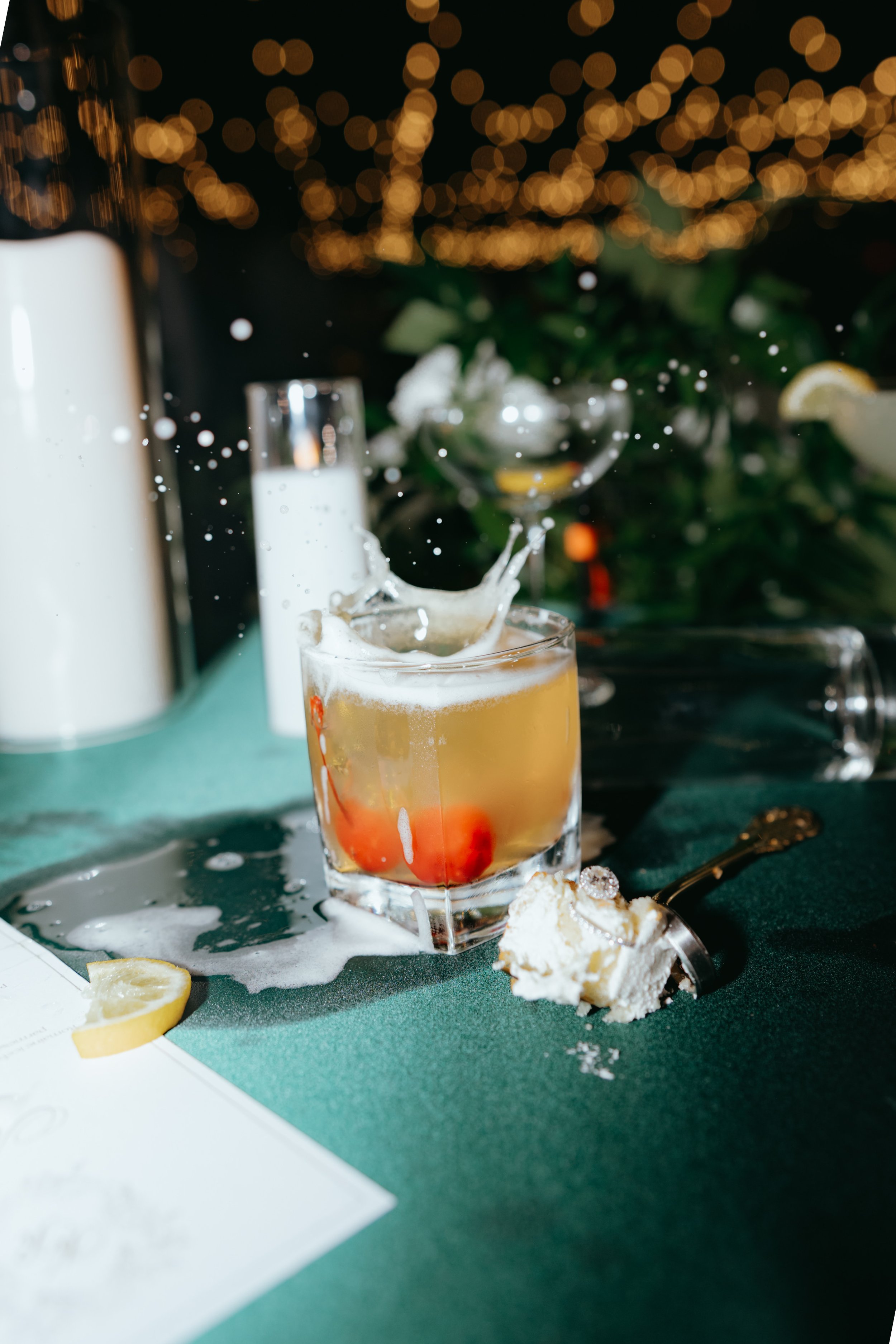 A splash in a glass of cocktail with cherries, lemons, and ice; a cake slice on a spoon nearby; spilled foam and lemon wedge on table; blurred background of string lights and greenery.
