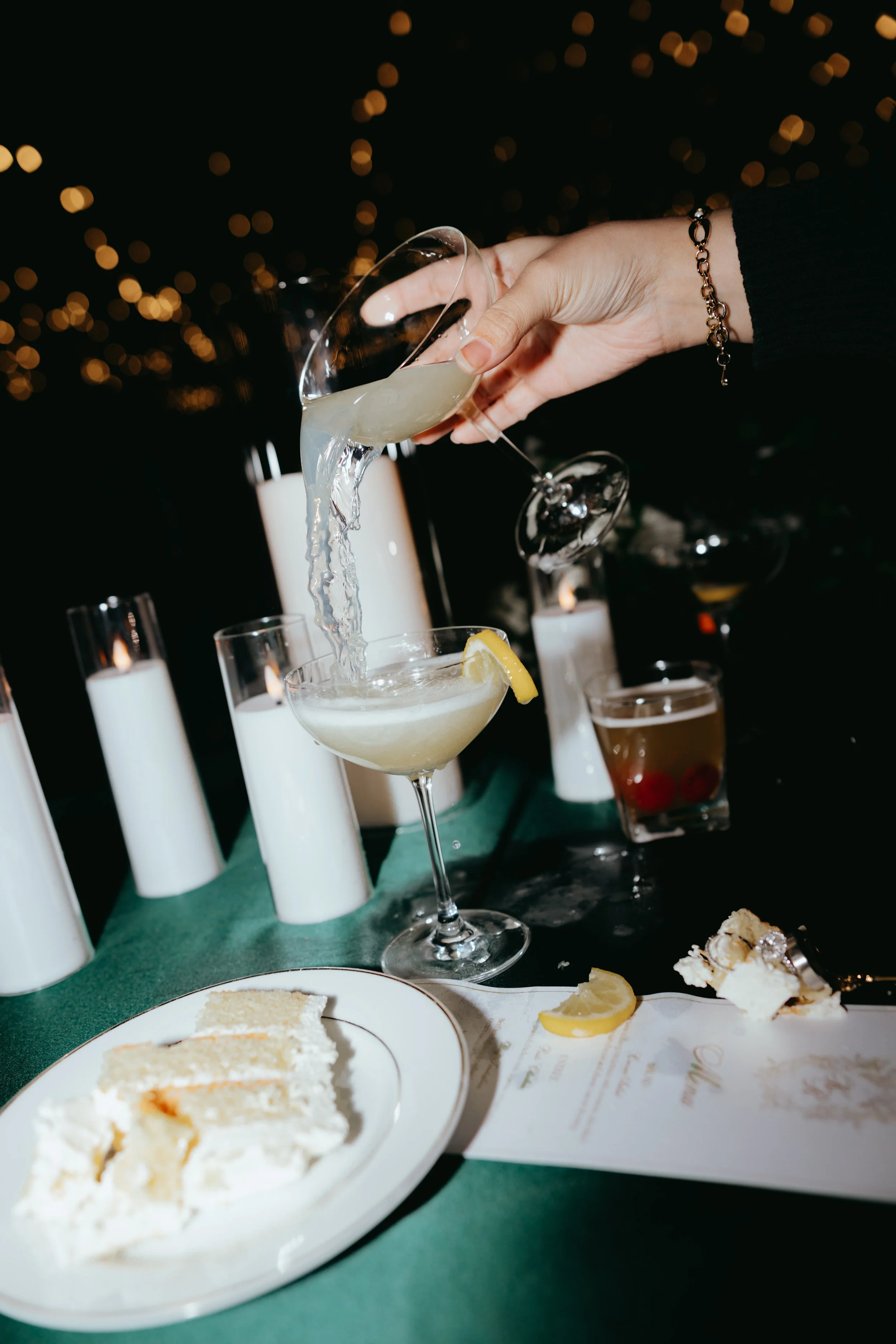 A person pours a clear cocktail into a coupe glass garnished with a lemon wedge, with candles, a plate of cake, and other drinks on the dark table.