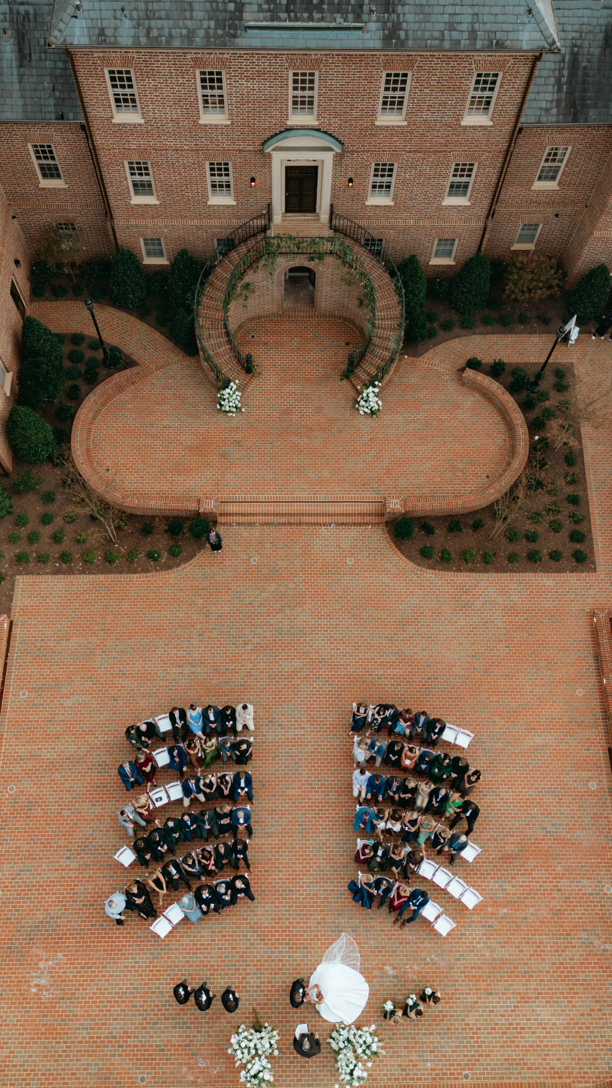 An aerial view of a wedding ceremony with guests seated in rows, a bride in a white dress, a groom, and officiants at the front on a brick-paved courtyard, surrounded by a brick building with a landscaped area and decorative lighting.
