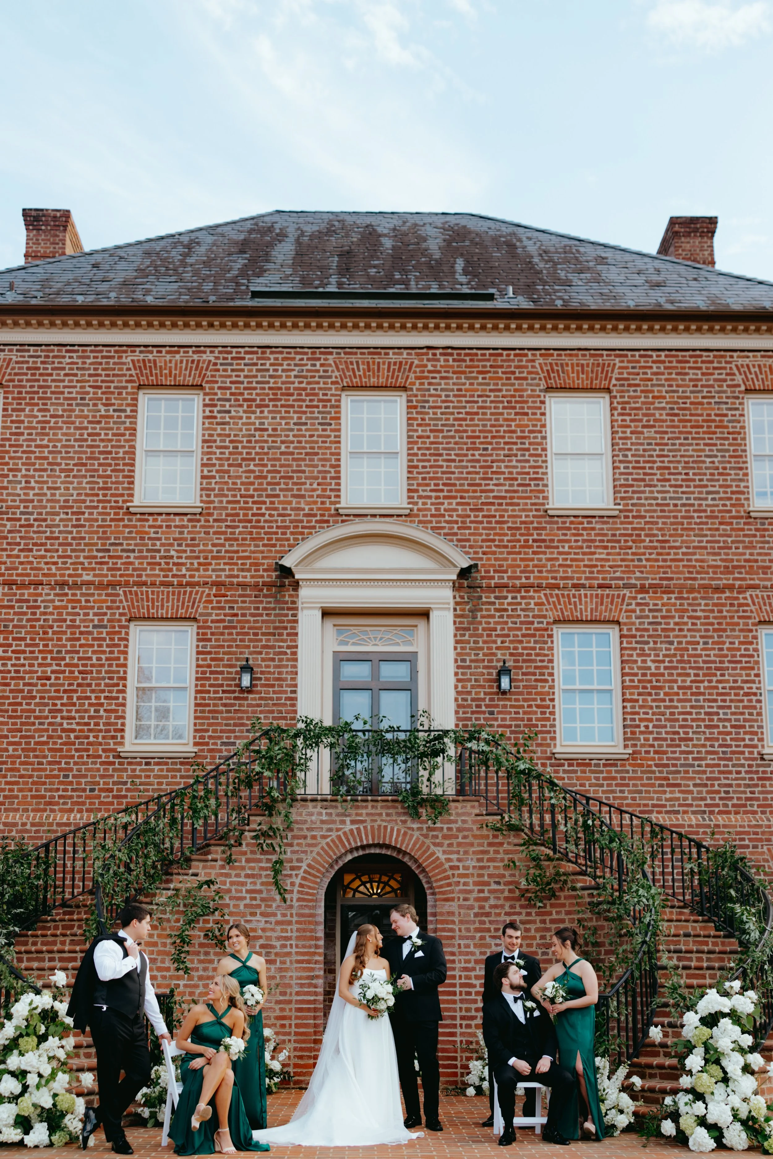 A wedding party arranged on a brick staircase outside a red brick building with six windows and decorative trim. The bride and groom are in the center, surrounded by bridesmaids and groomsmen, all dressed formally with floral arrangements.