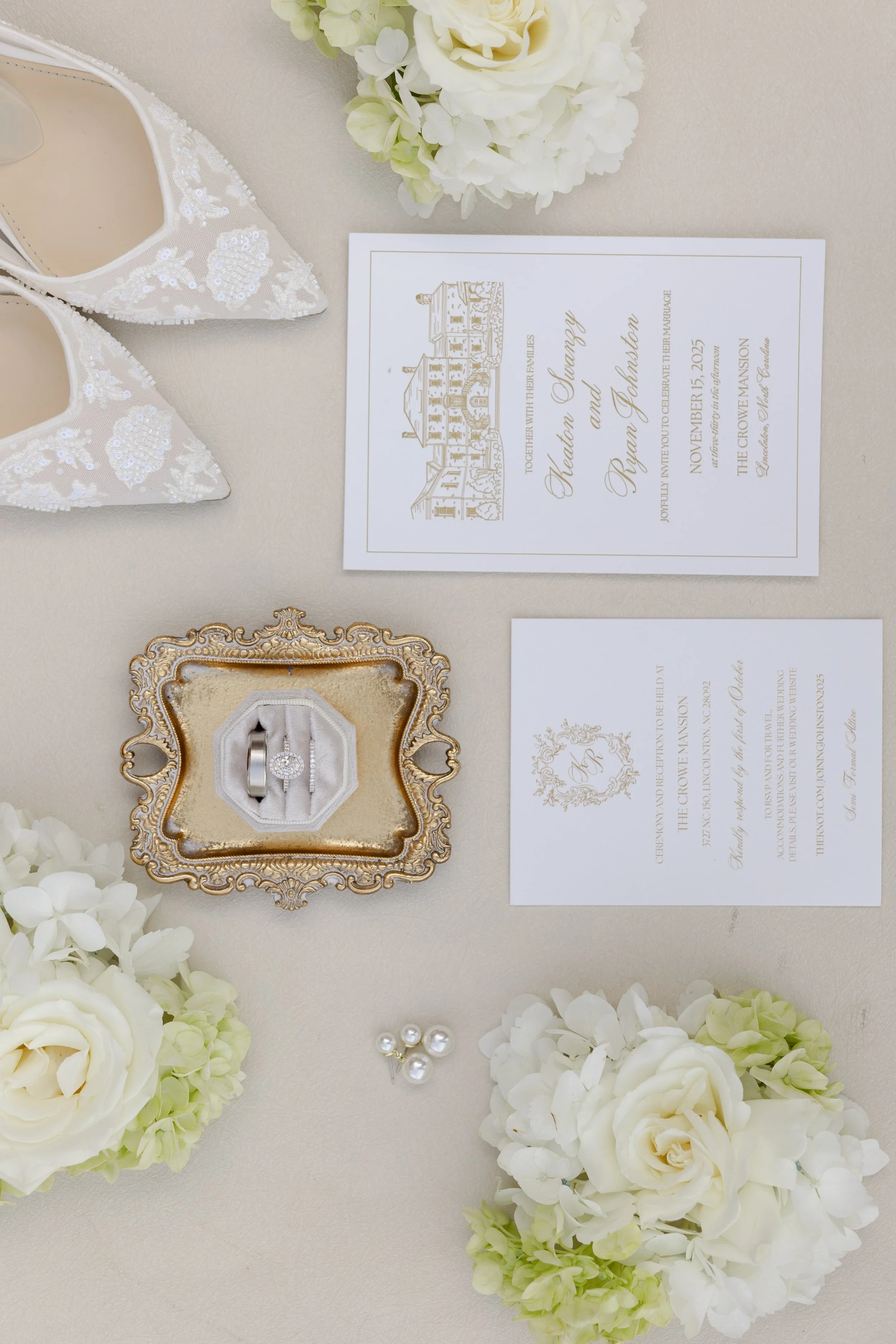 Wedding invitation, white shoes with beadwork, white flowers, and a wedding ring in a decorative gold tray.