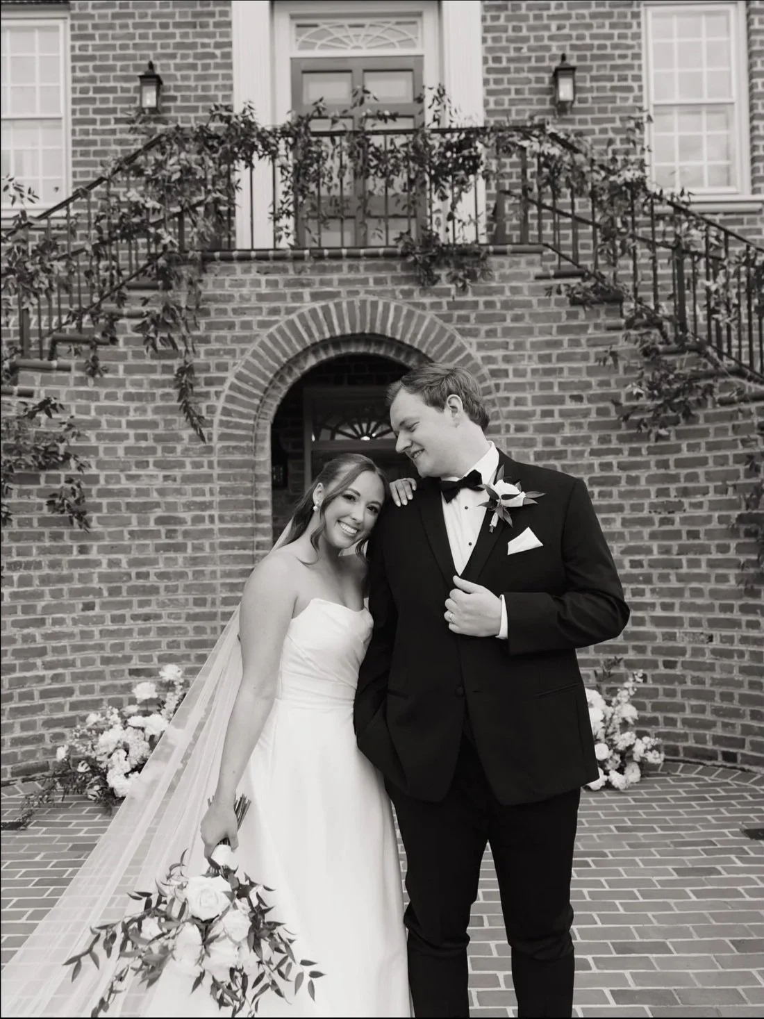 A black-and-white photo of a bride and groom standing outdoors in front of a brick building, smiling and looking at each other. The bride holds a bouquet of flowers, and the groom is dressed in a tuxedo with a boutonniere.