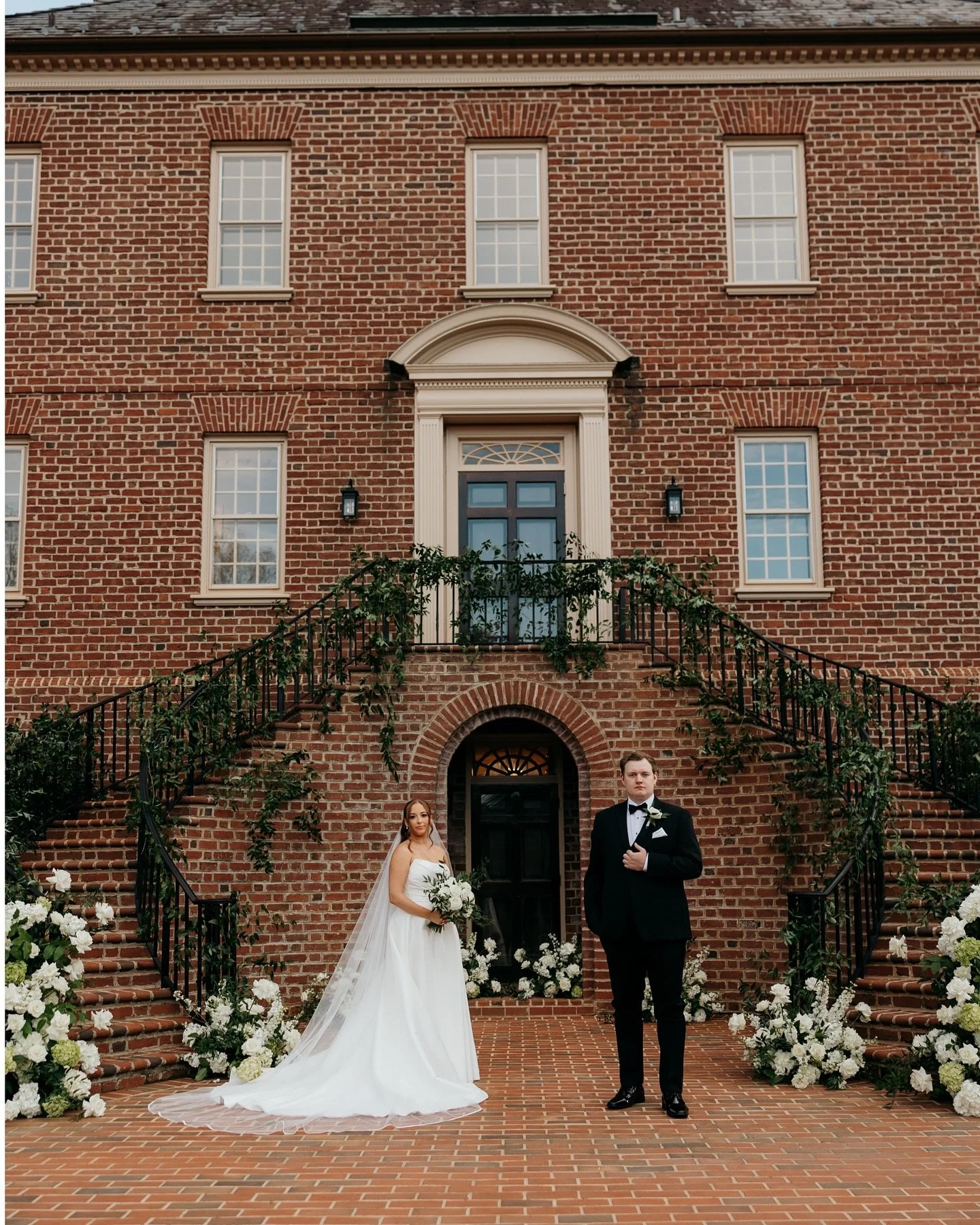 A bride and groom standing on brick steps outside a large brick building decorated with white flowers and greenery for a wedding.