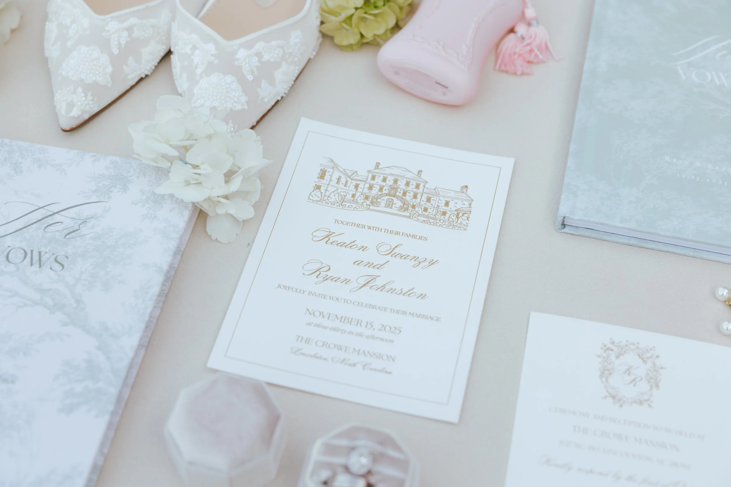 Wedding invitation with floral decorations, a pink perfume bottle, a lace handkerchief, and jewelry on a table.