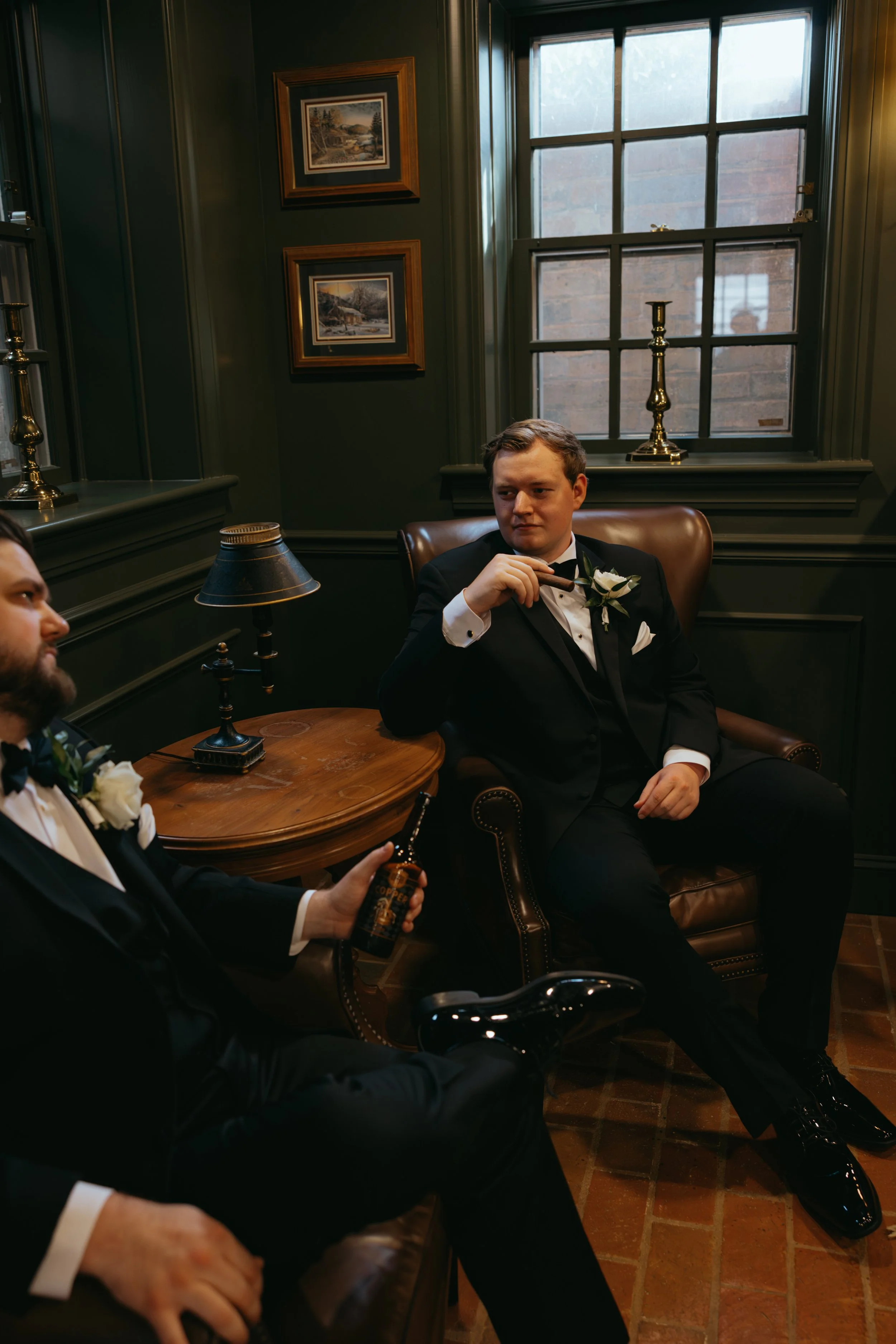 Two men in tuxedos sit in a vintage-style room with dark green walls, a large window, framed landscape paintings, and brass candlesticks. One man holds a bottle, the other sits relaxed in a leather armchair, holding a cigar.