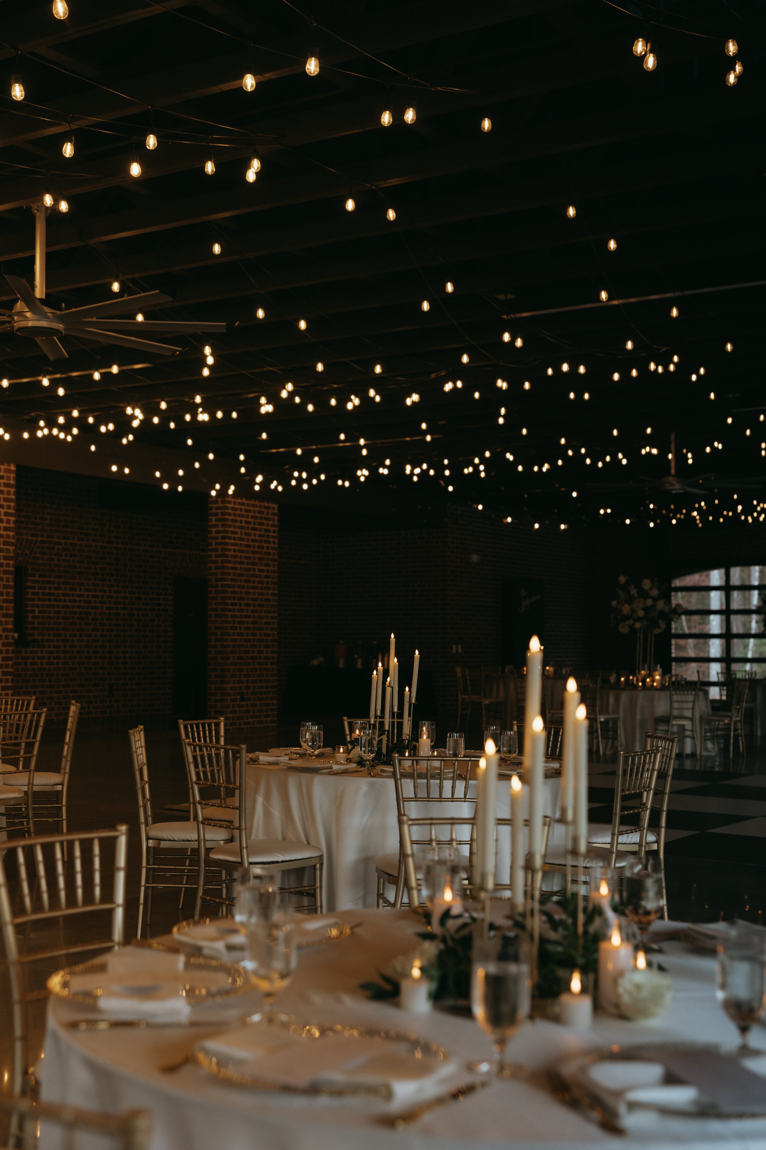 Elegant banquet hall decorated with string lights hanging from the ceiling and tall candles on tables, set for a formal event such as a wedding reception.