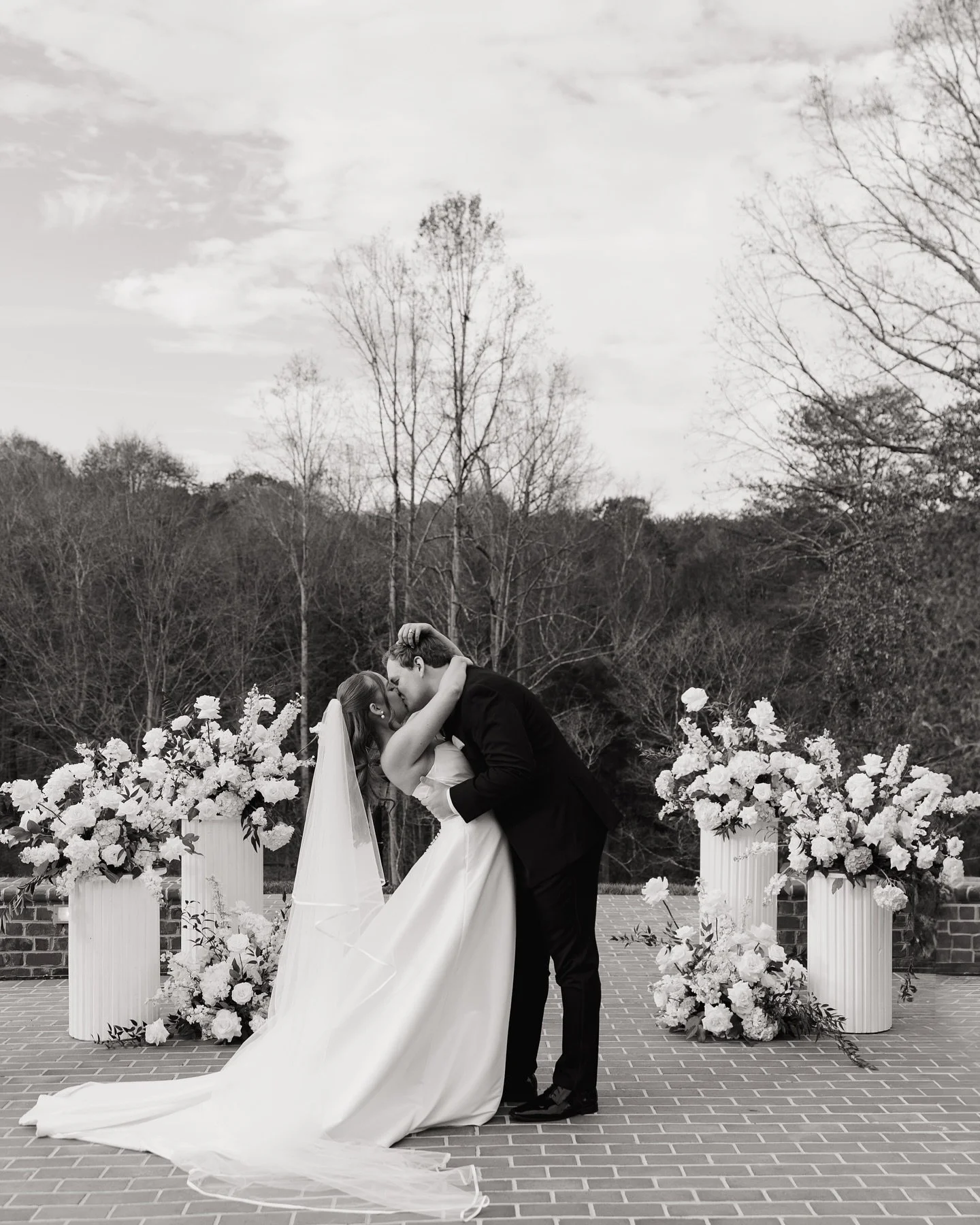 A black and white photo of a bride and groom kissing during their wedding ceremony outdoors, with floral arrangements on pedestals behind them.