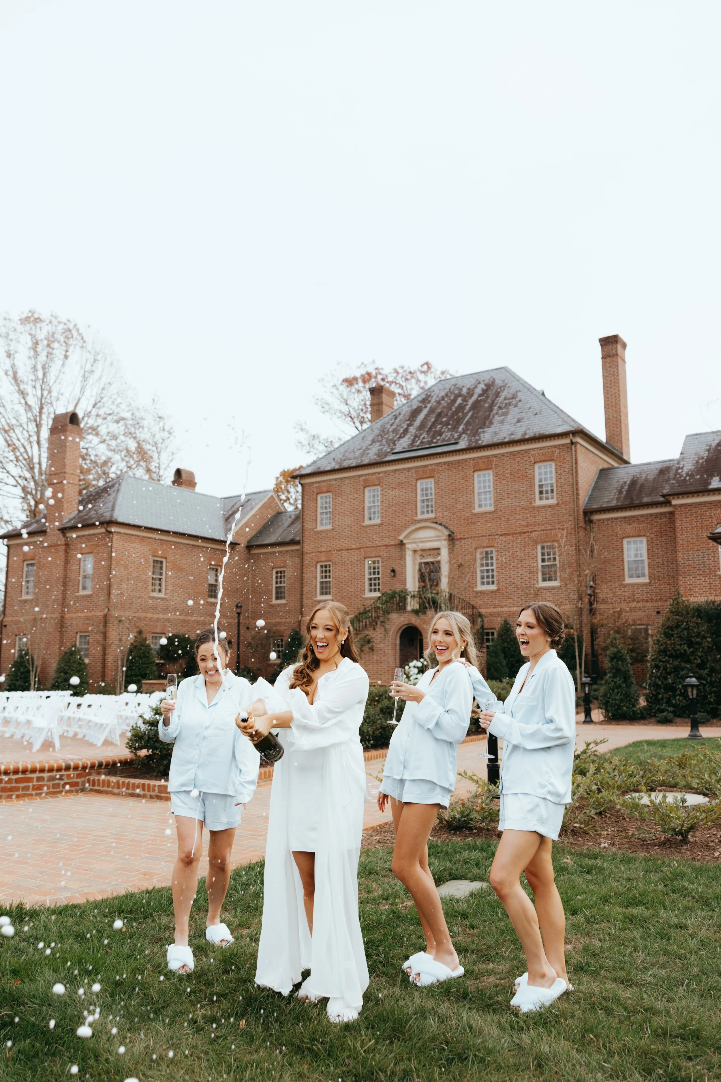 A group of four women in pajamas celebrating with a champagne bottle and falling foam outside a large brick mansion.