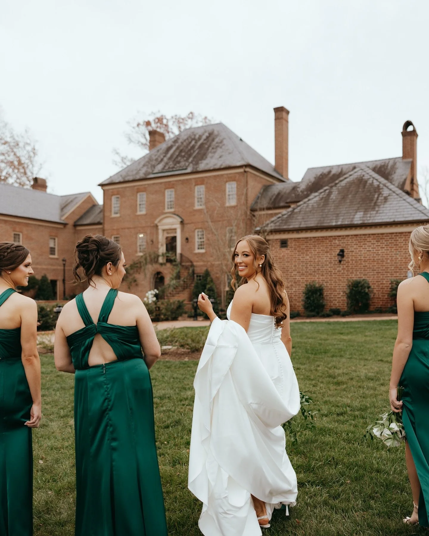 Bridal party standing on a lawn in front of a brick mansion, with the bride smiling and holding up her dress, surrounded by bridesmaids in dark green dresses holding bouquets.