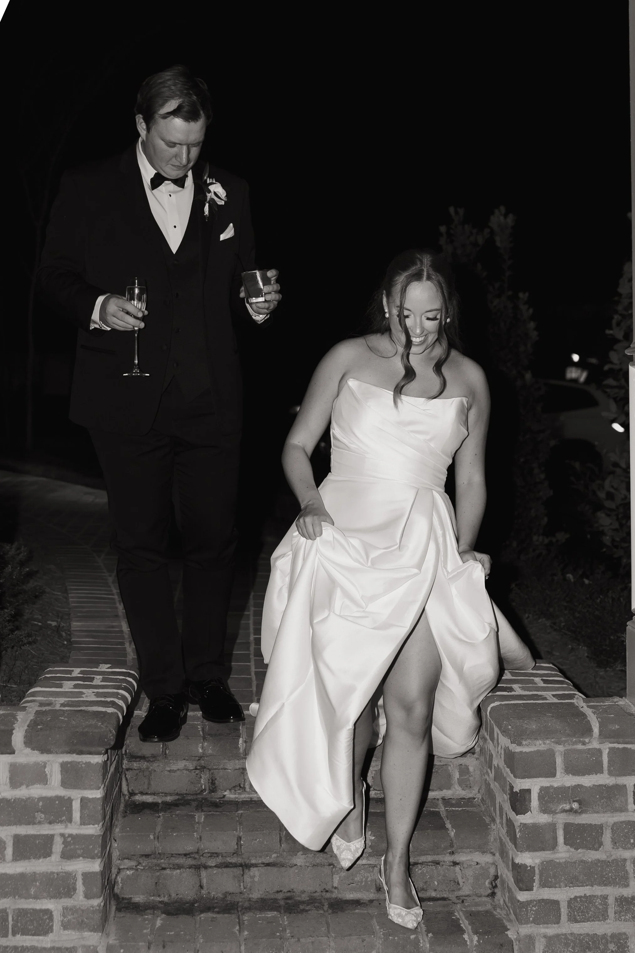 A bride in a strapless wedding dress and high heels is descending brick stairs, holding up her dress, with a cheerful expression. A man in a tuxedo with a flower boutonnière and holding a glass of champagne is behind her, looking down.