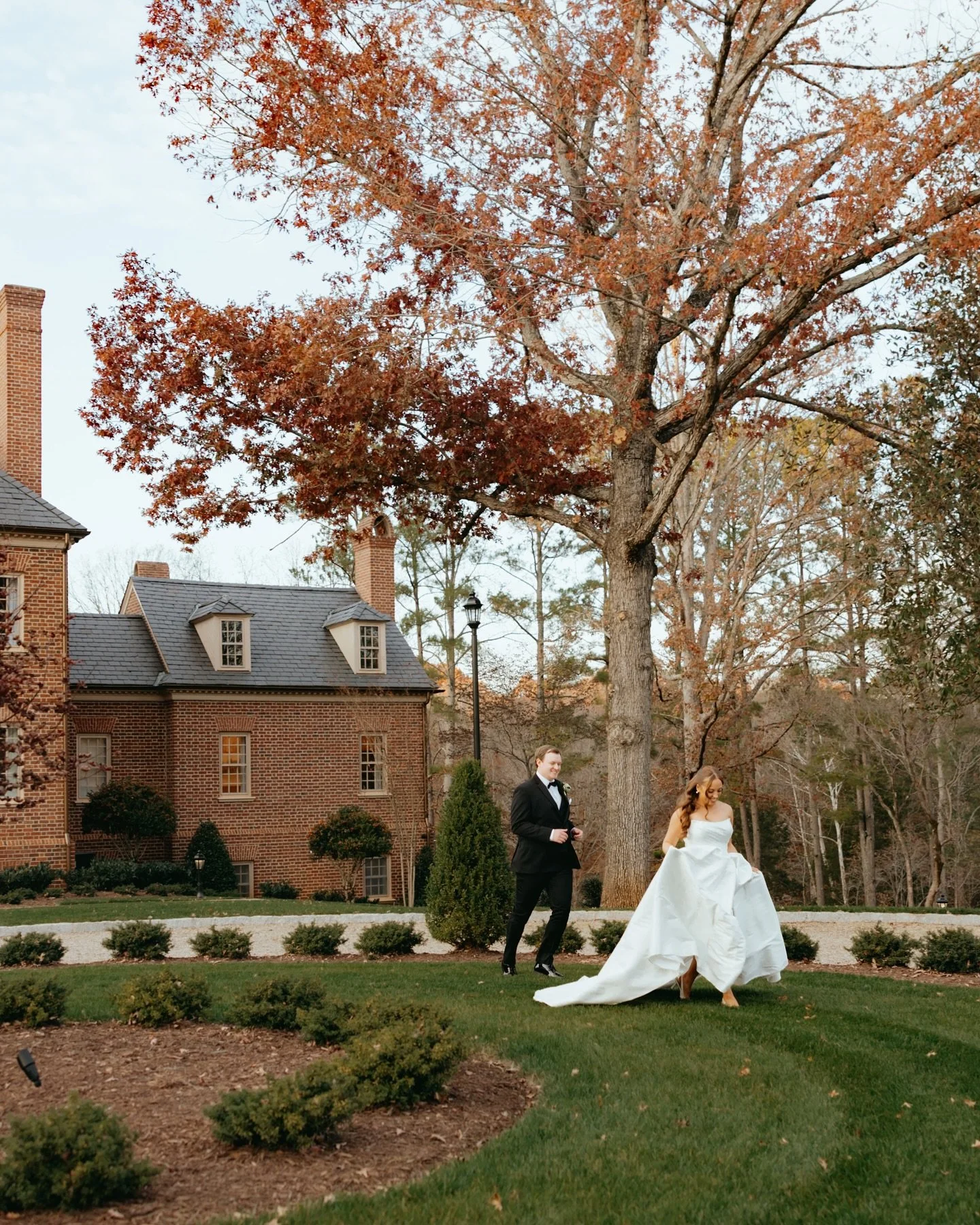 A bride in a white wedding gown and a groom in a black tuxedo walking on a grassy area with trees and a brick house in the background.