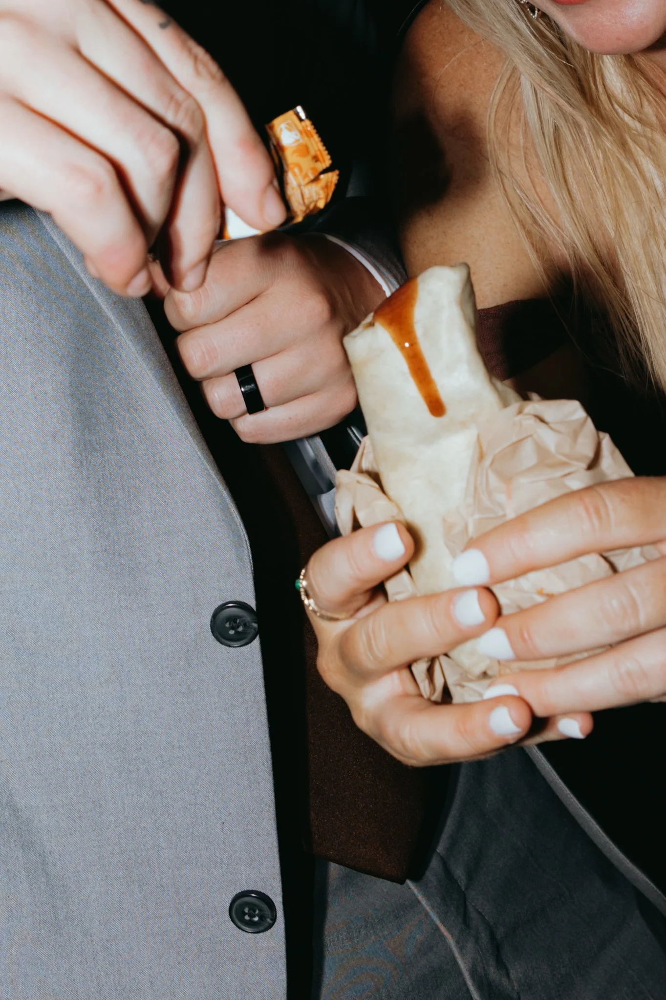 Person holding a burrito with white sauce and caramel drizzle, wrapping paper partially visible, person wearing a gray blazer and white shirt underneath.