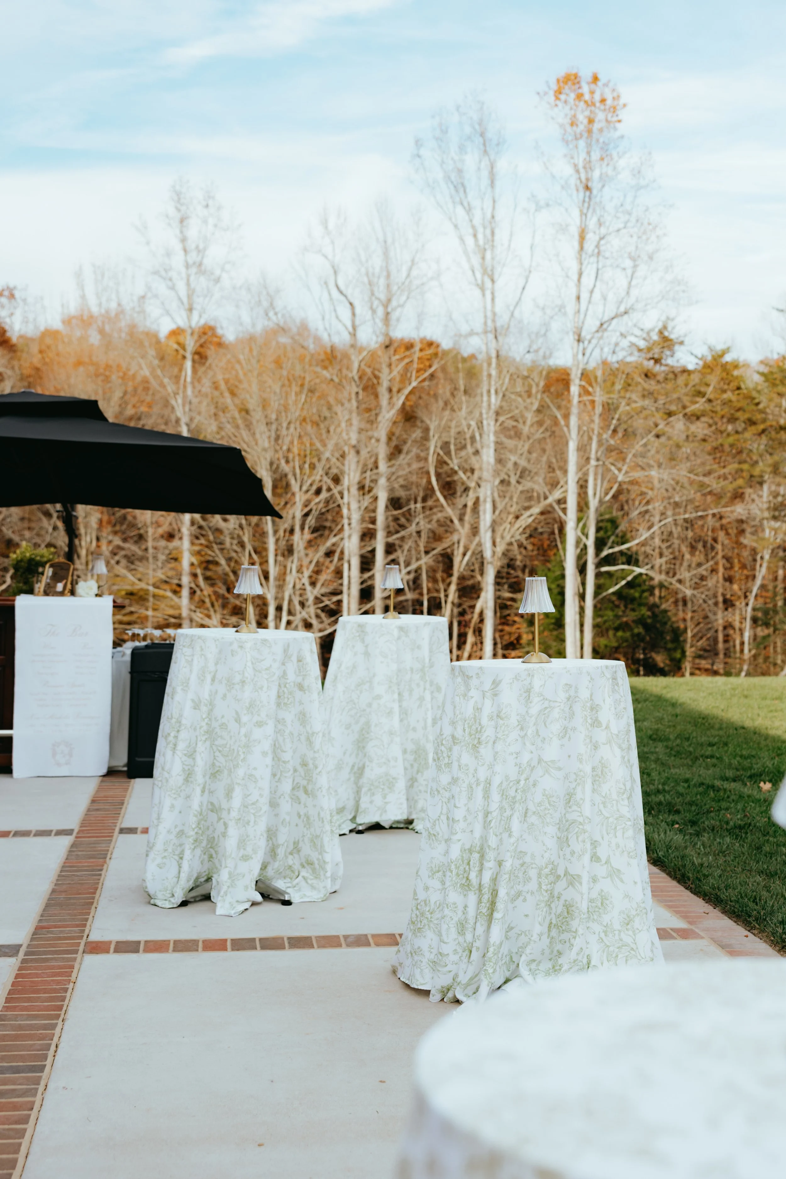 Outdoor wedding reception setup with draped tables, small lamps, a black umbrella, and a backdrop of fall trees.