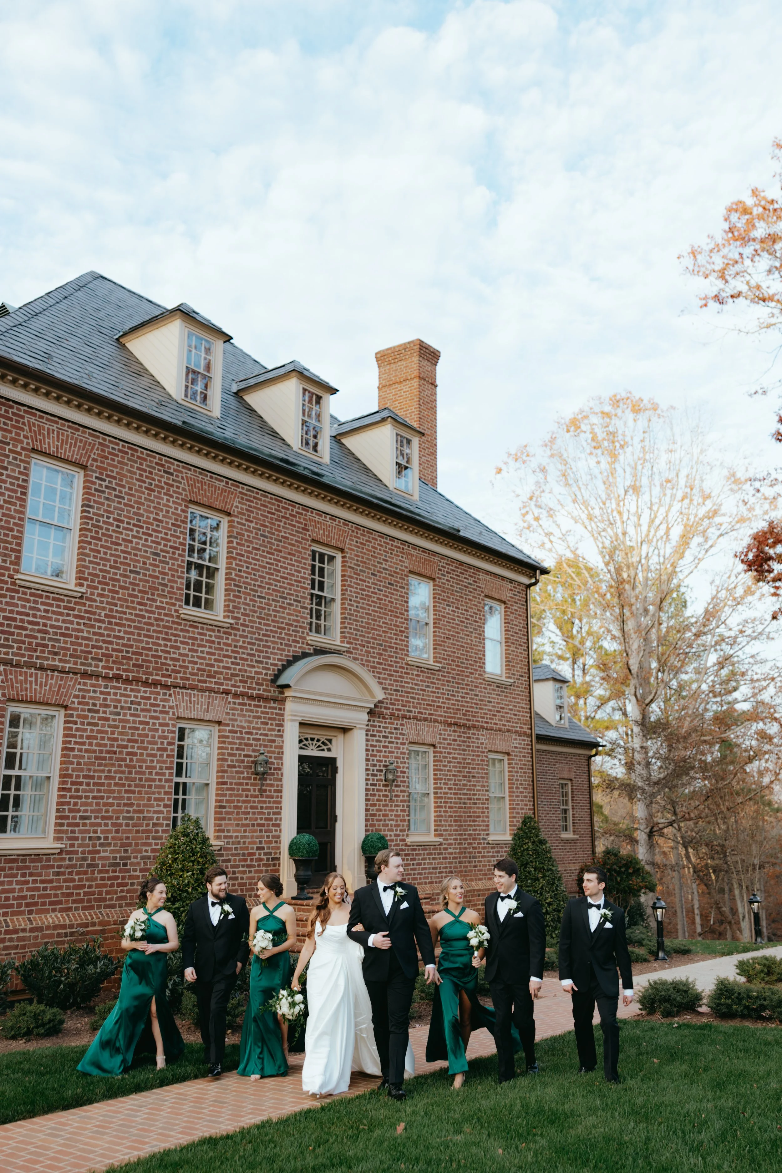 A wedding party walking on a brick pathway in front of a large brick house with a dark roof, surrounded by autumn trees and grass.
