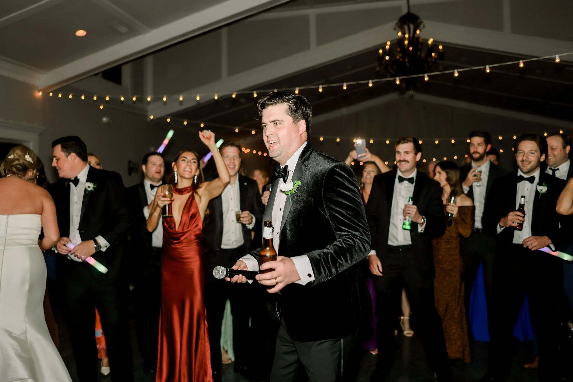 A wedding reception with a groom in a black tuxedo holding a microphone and a beer, surrounded by guests in formal attire, dancing and celebrating under string lights and a chandelier.