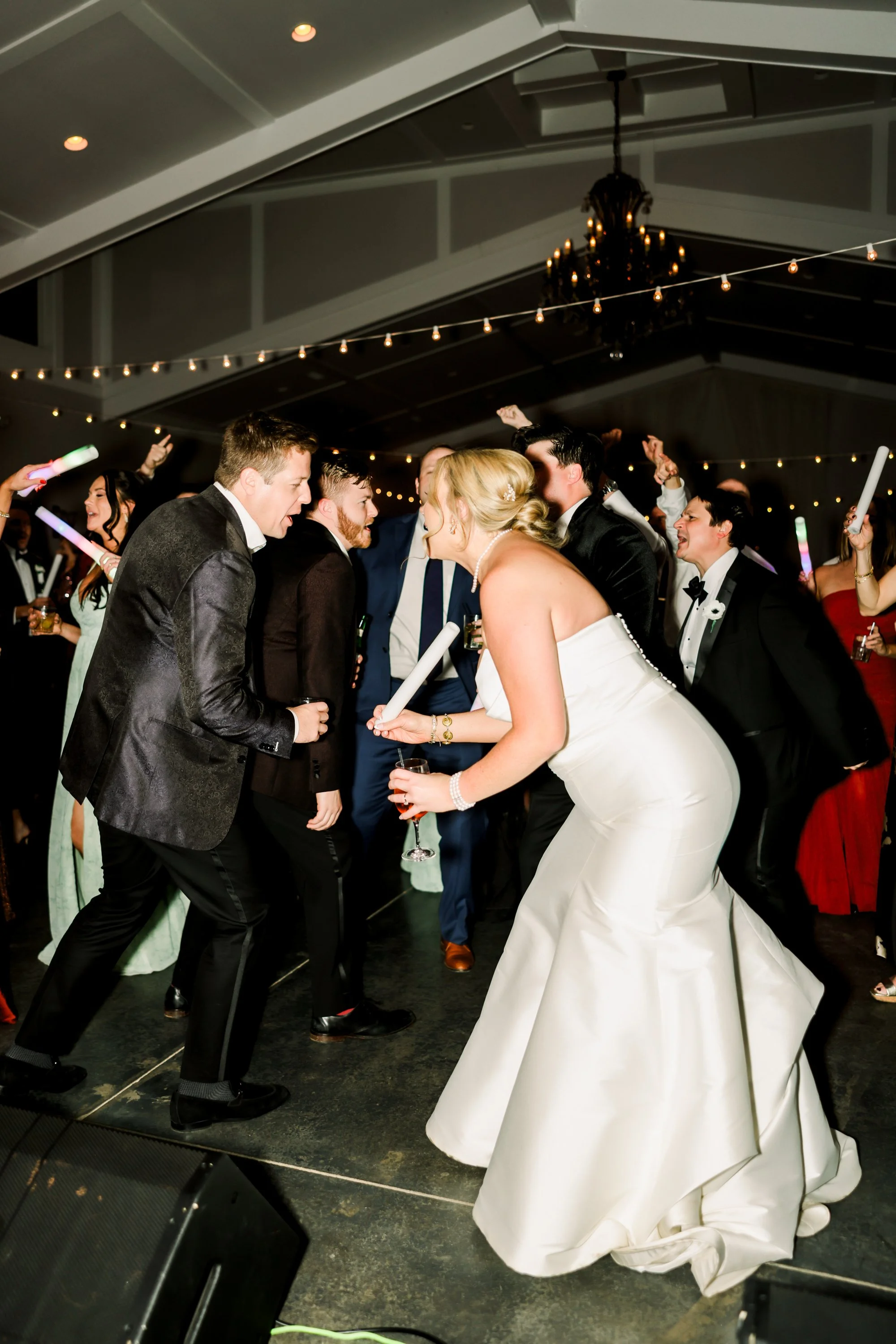 Bride and groom dancing at wedding reception with guests, holding glow sticks and drinks, under string lights and chandelier.