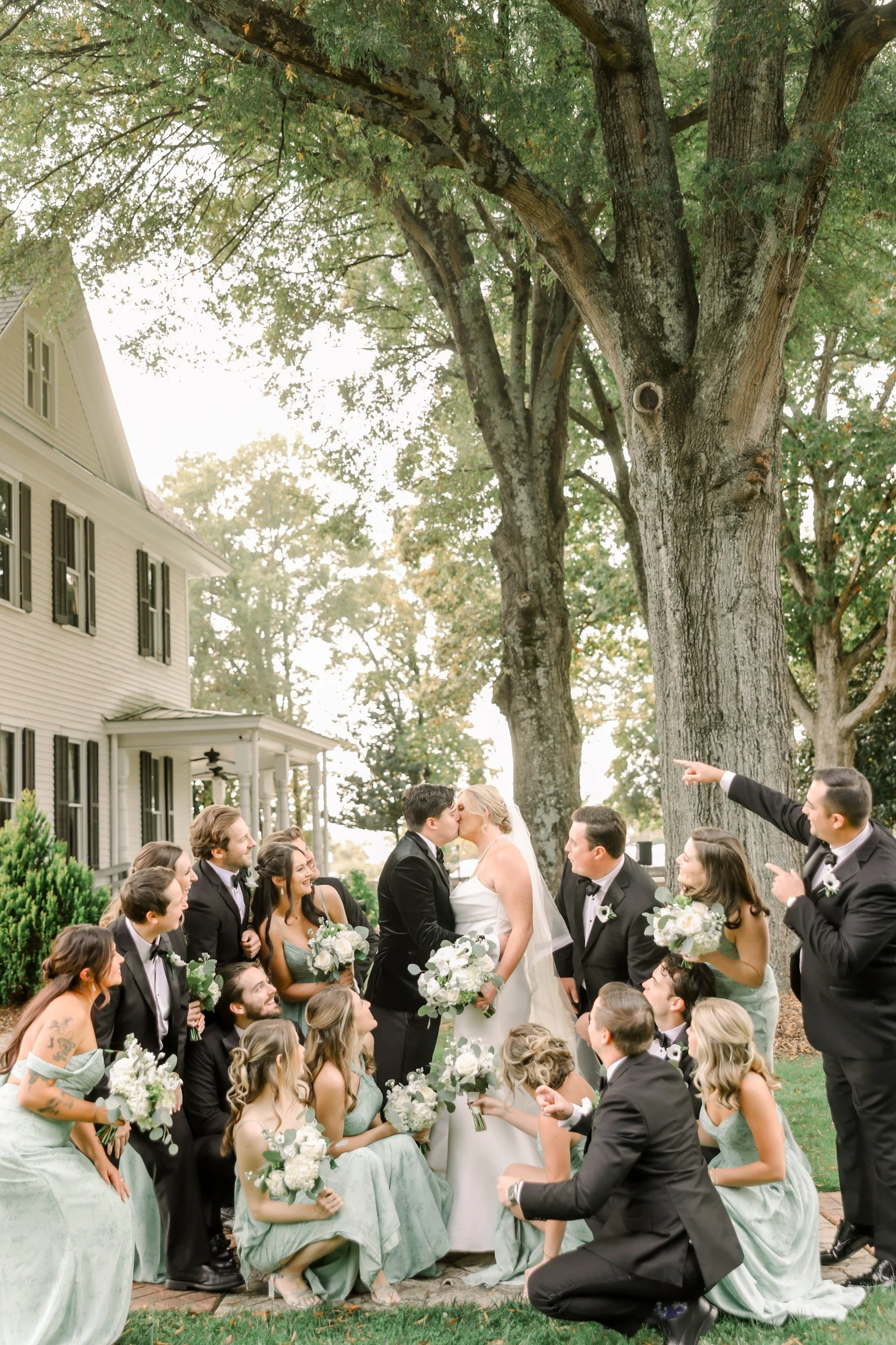 A wedding party outside, with the bride and groom kissing beneath large trees and surrounded by friends and family, some of whom are pointing or kneeling, holding bouquets and dressed in formal attire.