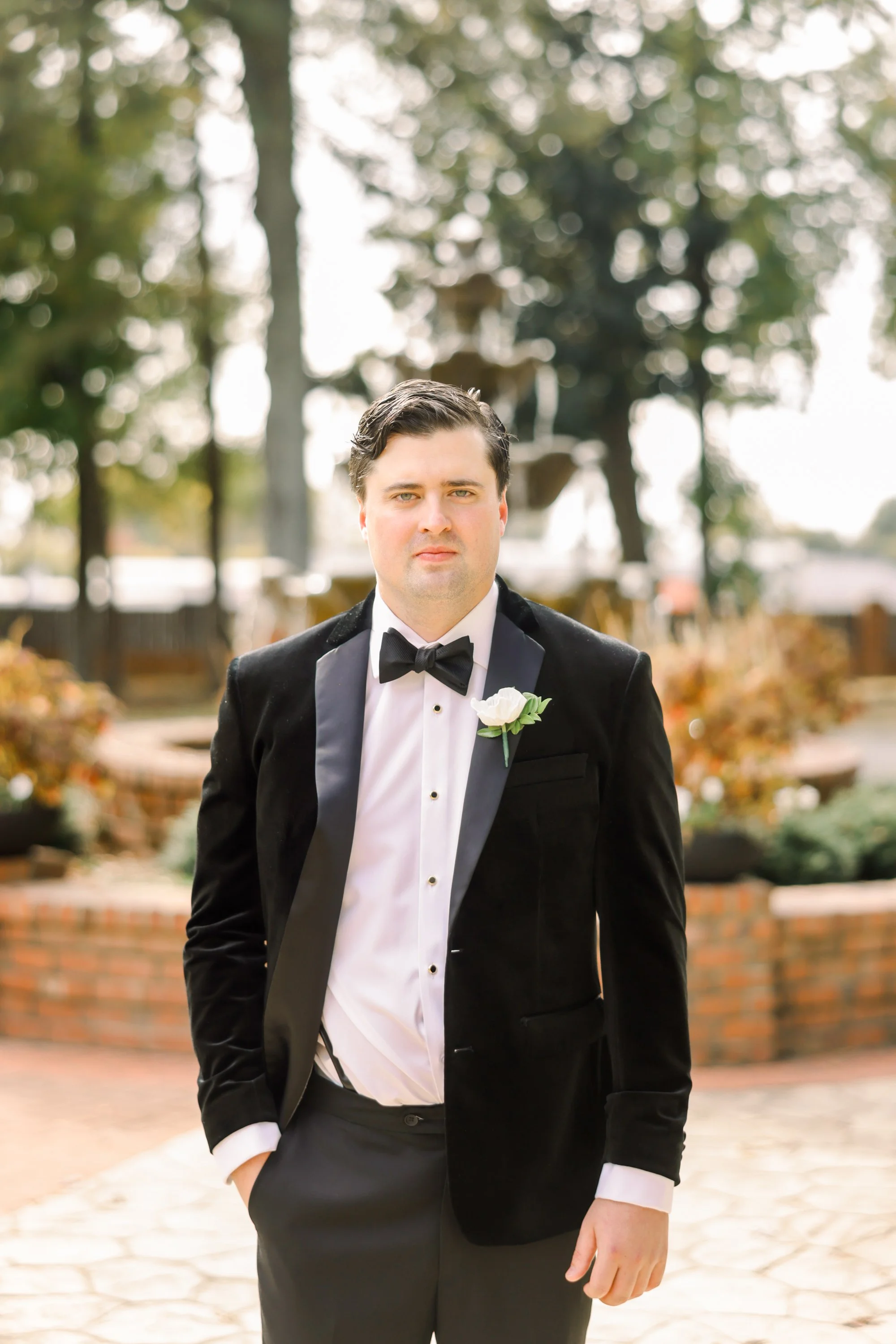 A man in a tuxedo with a white boutonniere standing outdoors in front of a fountain, trees, and brick structures.