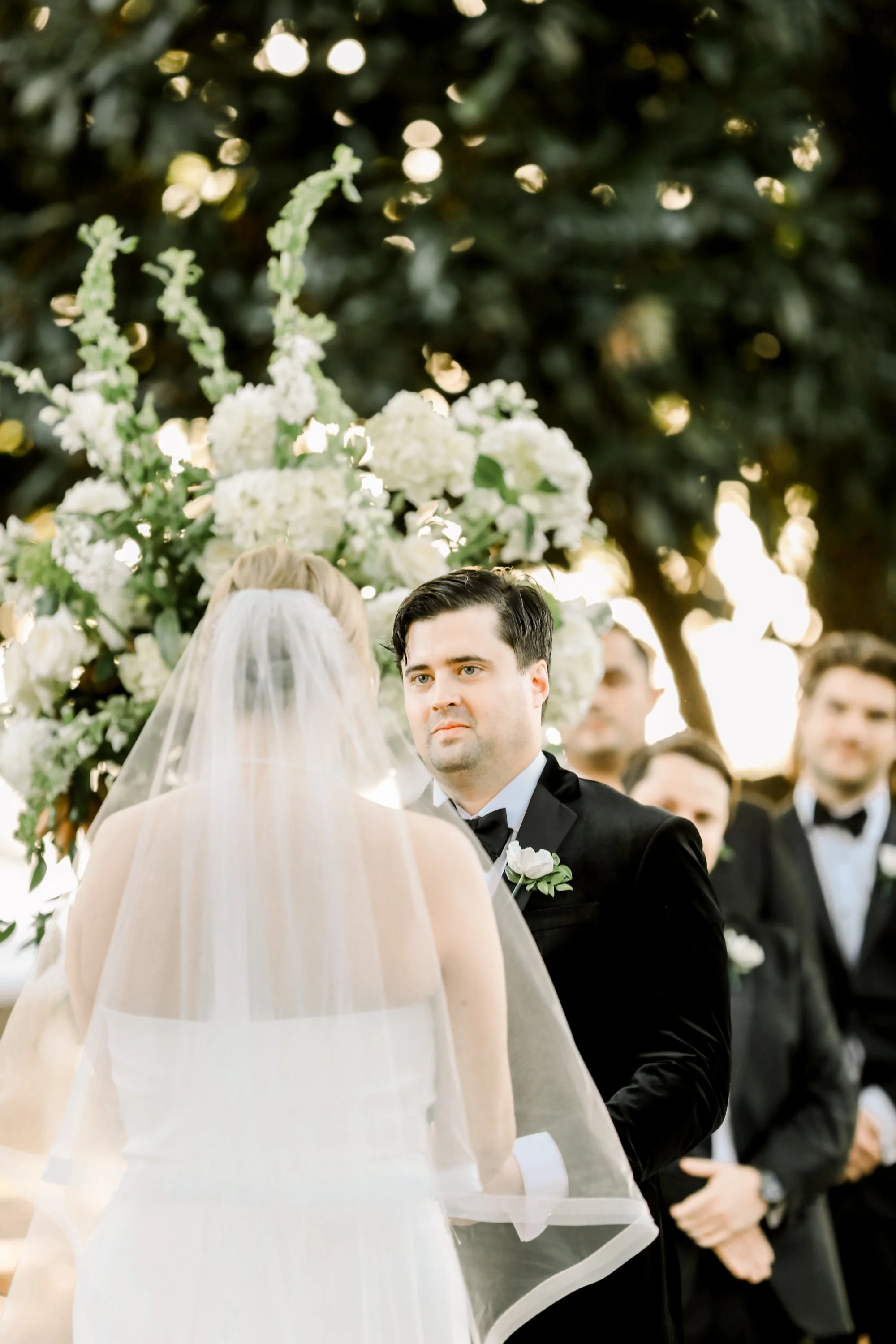 A groom in a black tuxedo looks at a bride in a white wedding dress and veil during their outdoor wedding ceremony, with a floral backdrop and wedding party in the background.