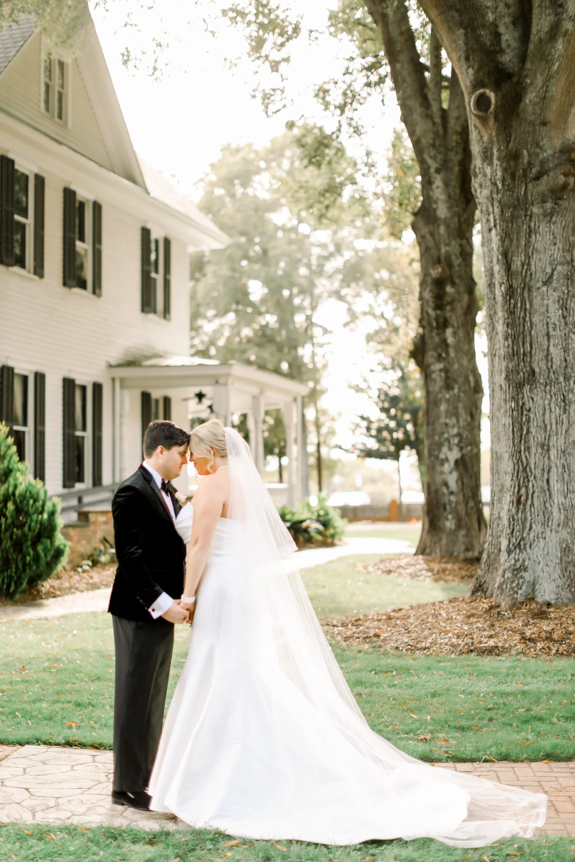 A bride and groom holding hands and touching foreheads outside near a house and large trees.