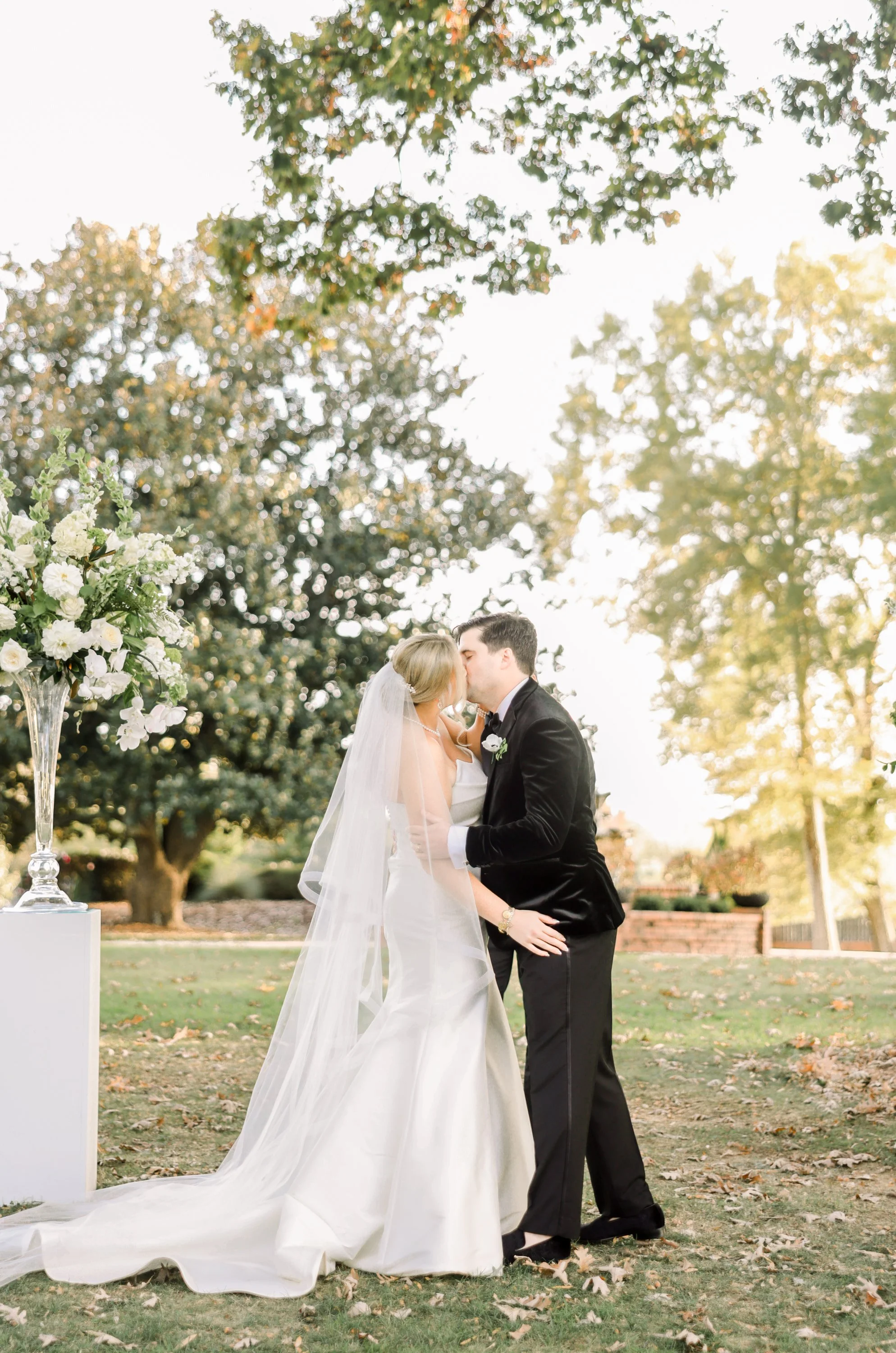 A bride and groom share a kiss during their outdoor wedding ceremony, with trees and sunlight in the background and a large floral arrangement on a white pedestal nearby.