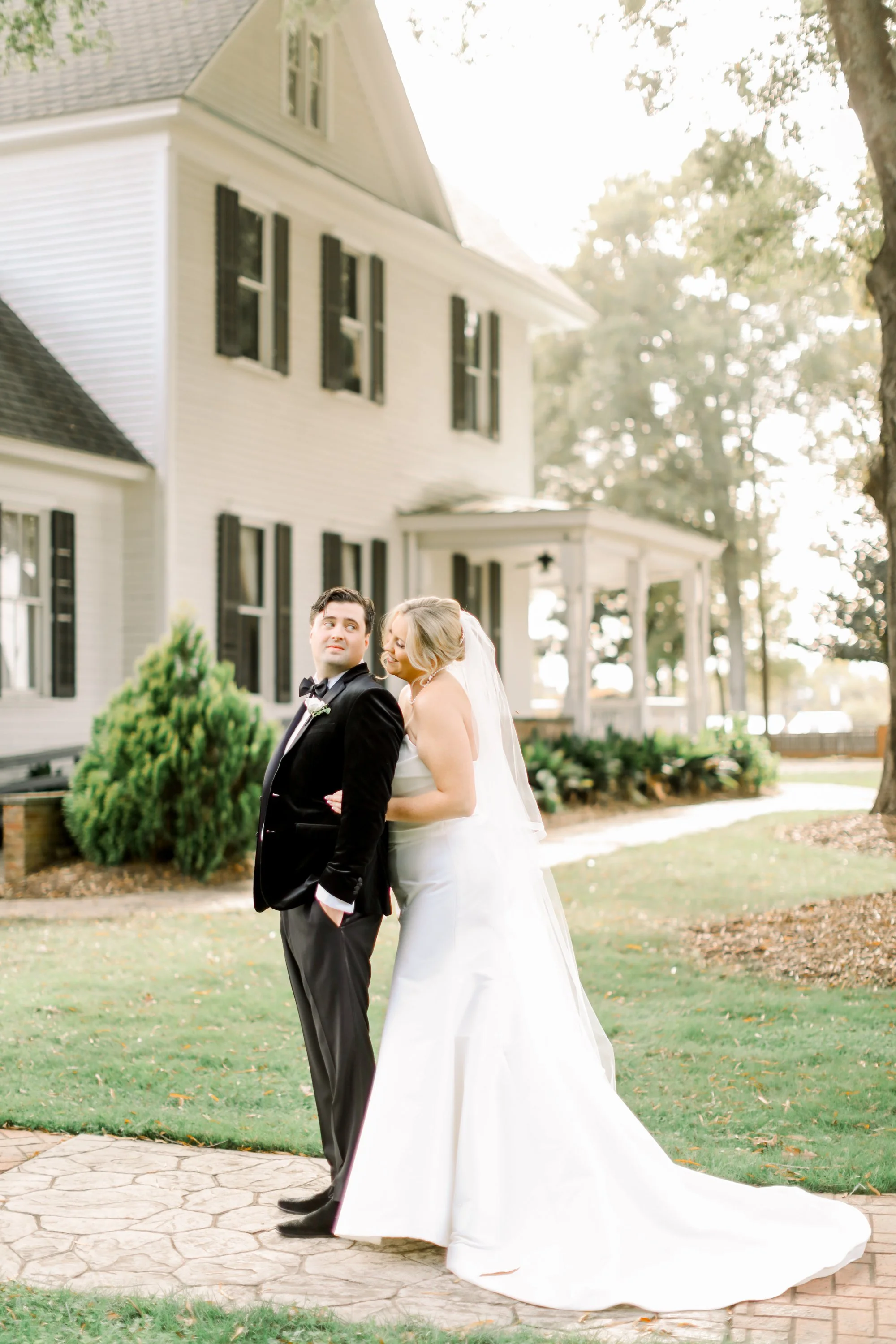 A bride and groom standing together outdoors in front of a white house, with the bride leaning into the groom.